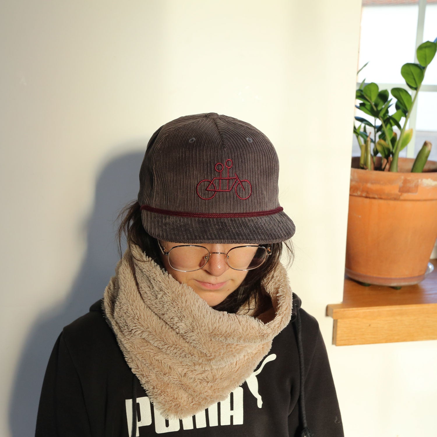 A person wearing a Tandem Coffee Roasters custom Corduroy Tandem Hat with a red bicycle logo, round glasses, and a large fluffy scarf, standing next to a potted plant in a sunlit room.