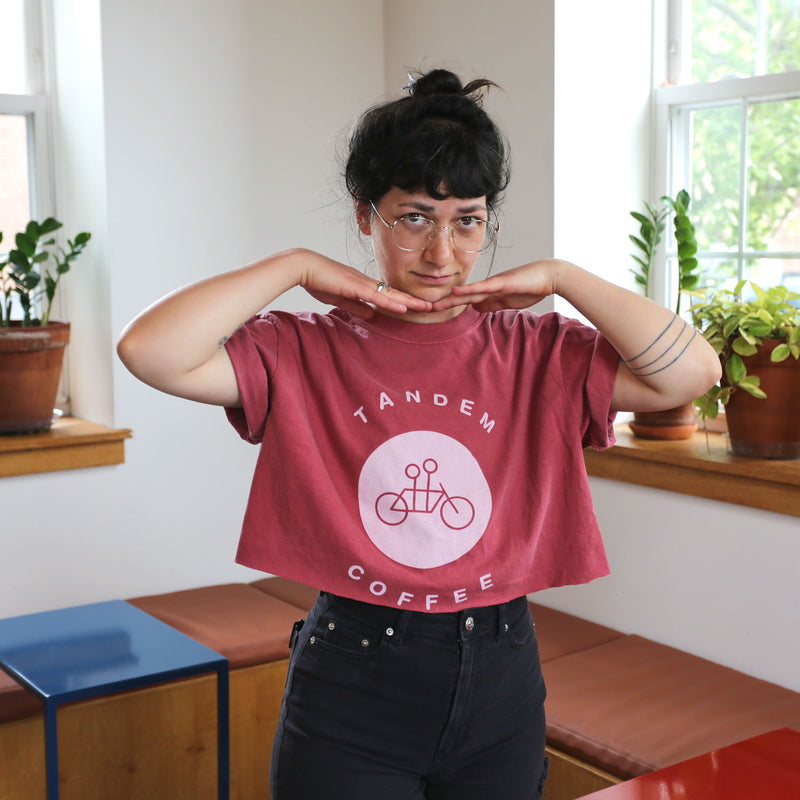 A person with glasses and a tattoo on their left forearm is smiling while sitting on a brown bench. They are wearing a Crimson Circle Logo Tee from Tandem Coffee Roasters with the text "Tandem Coffee" and an image of a tandem bicycle. A potted plant is on the windowsill behind them.