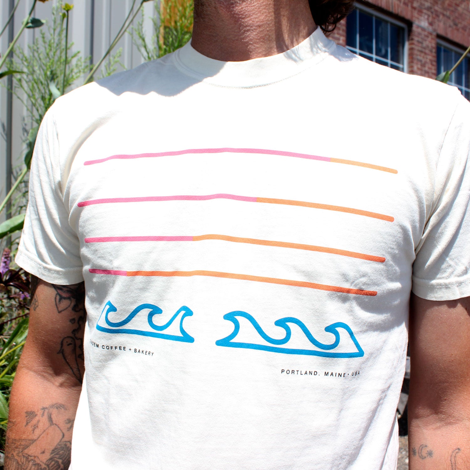 A person stands outside by a building with plants, wearing the Tandem Filthy Sunset Tee featuring wavy blue lines with orange and pink accents, giving off a neon art vibe. Limited stock available.
