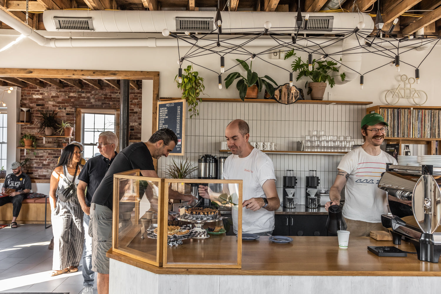 A cozy coffee shop in Portland, Maine, with exposed brick walls and ceiling beams. Two baristas in white shirts serve customers at the counter with a glass display of Tandem Coffee pastries. Other patrons wait in line, and plants and shelves with decor can be seen in the background.