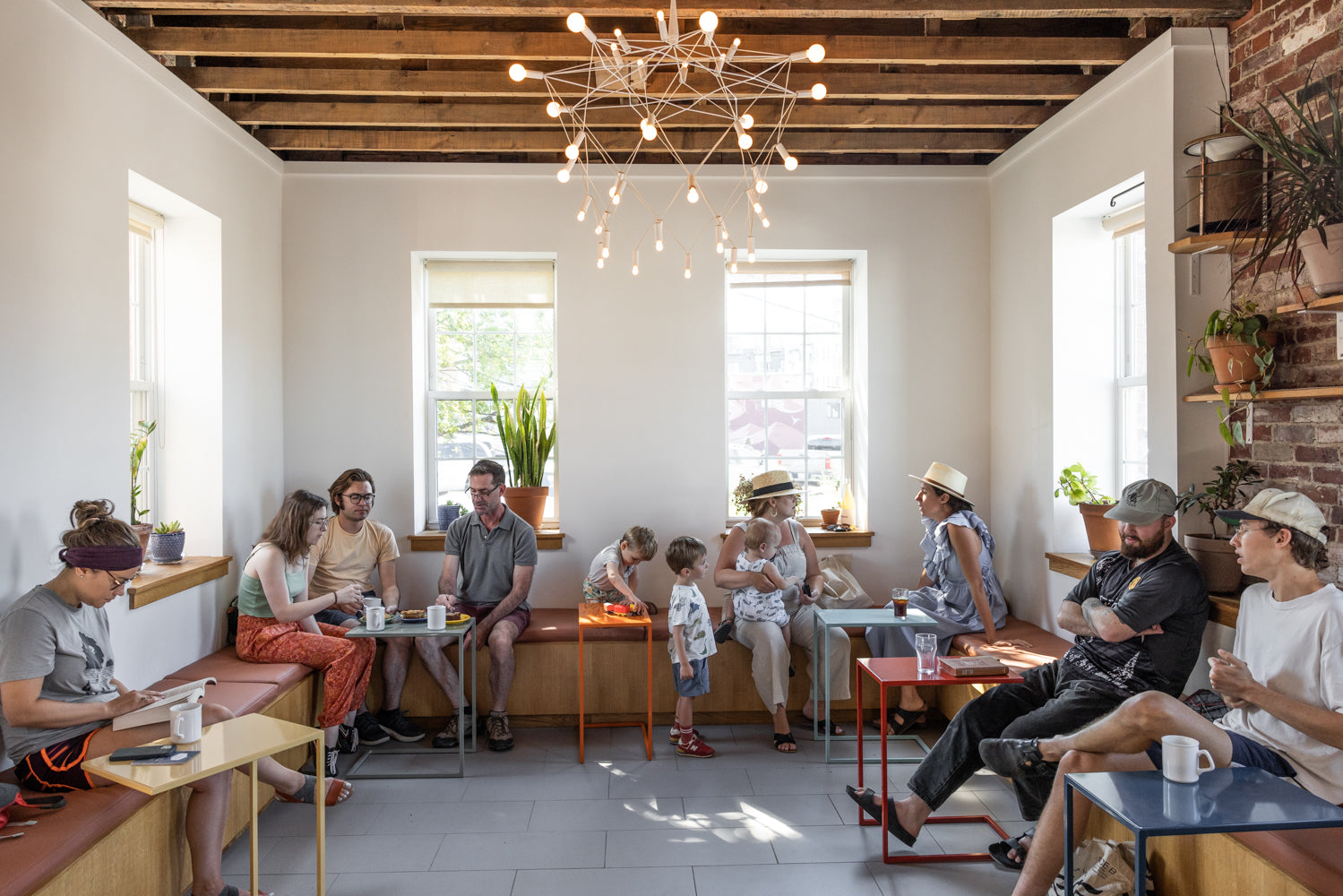 A casual, brightly-lit cafe in Portland, Maine features a modern, minimalistic design with white walls and wood accents. People of various ages are sitting, talking, and enjoying their tandem coffee around colorful tables. A unique light fixture hangs from the wooden ceiling, and large windows provide ample natural light.