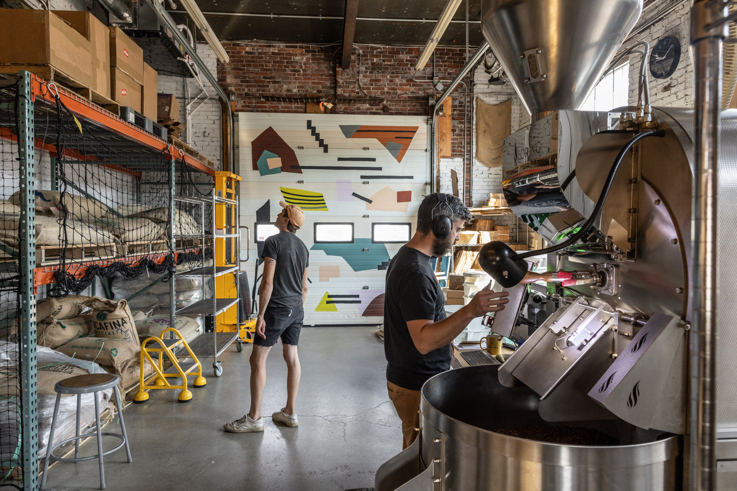 Two individuals work at Tandem Coffee in Portland, Maine. One person, wearing a cap and apron, handles a large coffee roasting machine, while another stands near shelves of coffee sacks, facing a garage door with colorful geometric designs. The setting has an industrial feel.