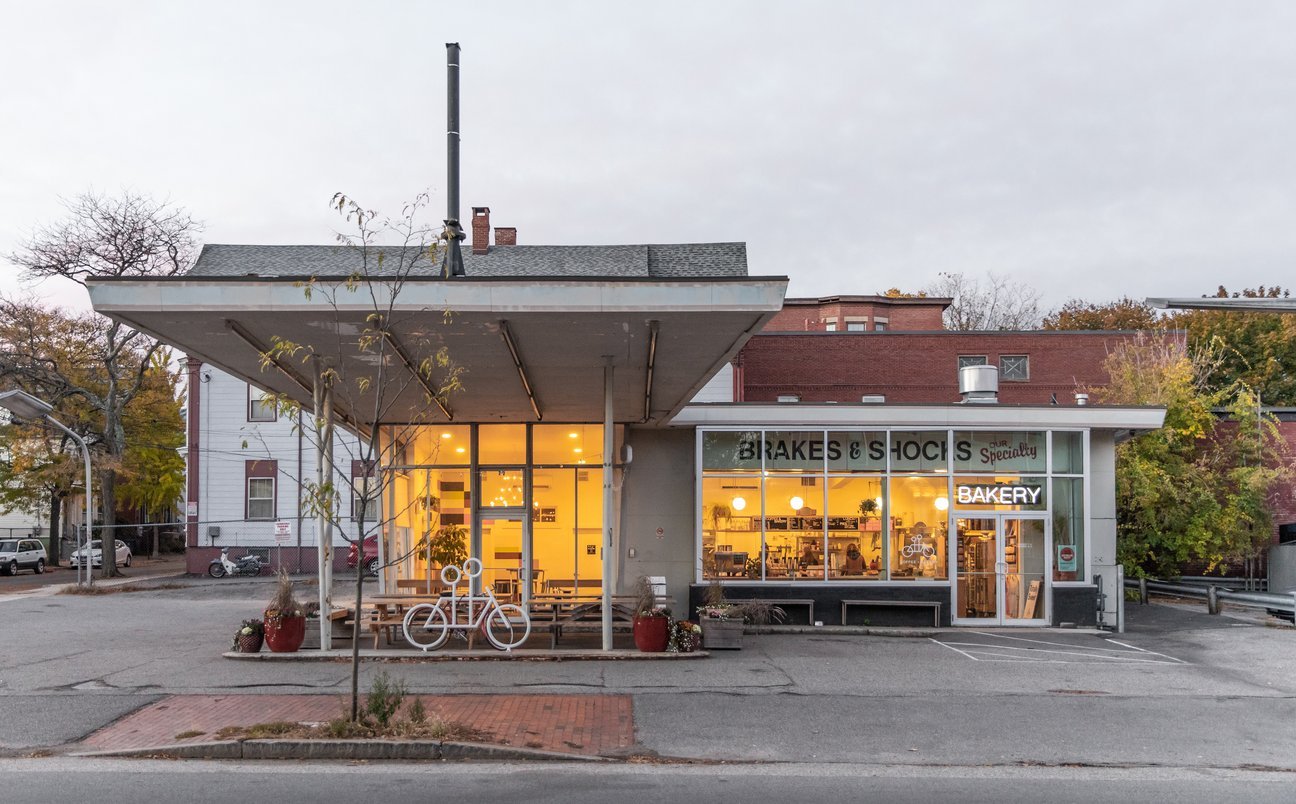 A quaint bakery, "blake's," housed in a unique building with a flat roof and large glass windows at dusk, displaying a warm, inviting interior. the street in front is quiet, with a bike parked outside.