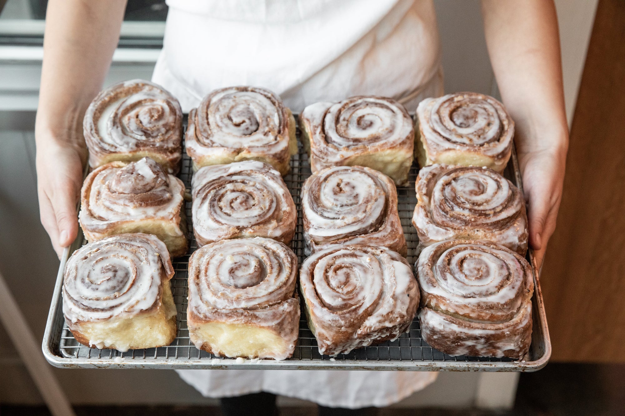 A person in a white apron holds a tray of freshly baked cinnamon rolls, each swirled with icing, displayed neatly within a metal baking tray.