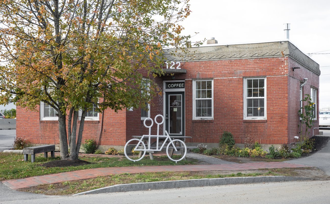 A small brick coffee shop with a white bicycle rack shaped like a bike in front, located at 122 street. a tree with autumn leaves and a bench are on the left.