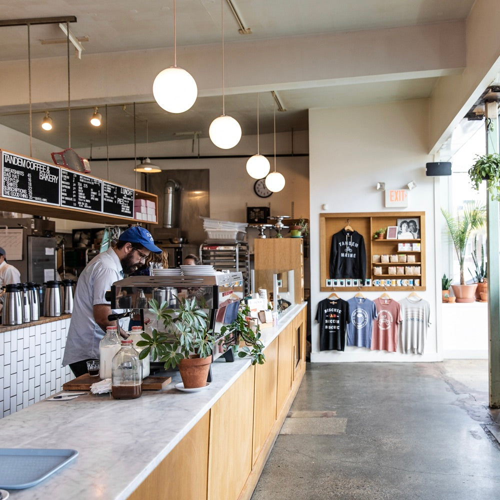A barista wearing a blue cap works behind the counter at Tandem Coffee in Portland, Maine. The counter is lined with plants and coffee-making equipment. A display on the right showcases merchandise including t-shirts. Hanging globes light up the bright and airy space.