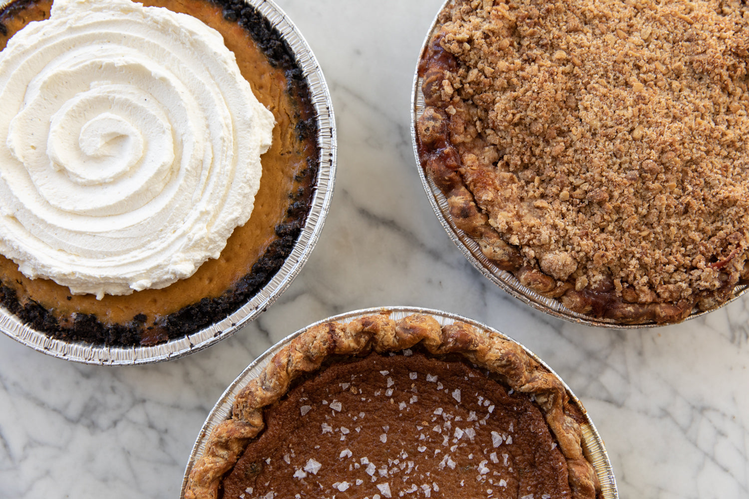 Three pies on a marble countertop from top view: a whipped cream topped pumpkin pie, a crumble-covered apple pie, and a chocolate pie with flaked salt.