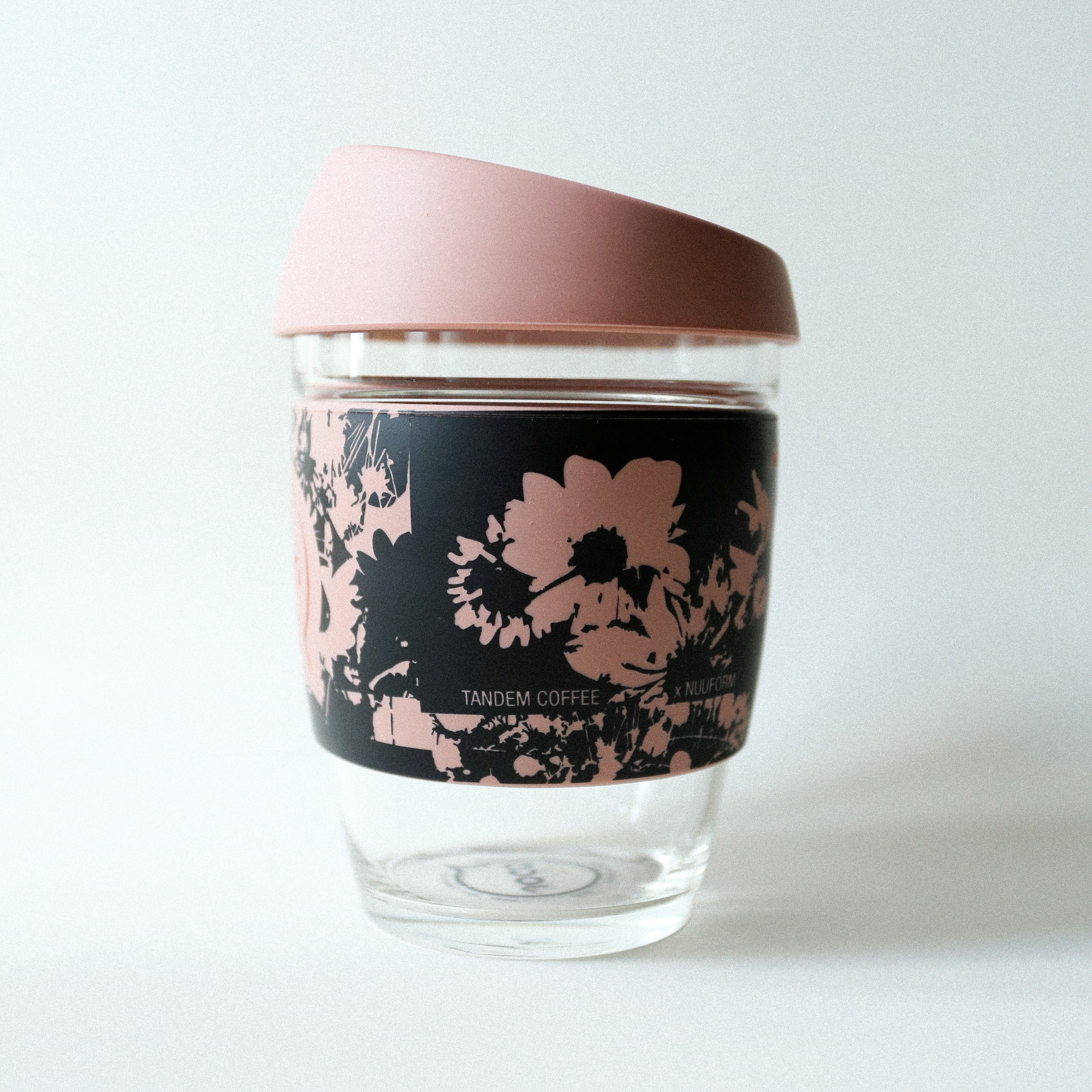 The Tandem x NUUFORM Joco Cup by vendor-unknown features clear borosilicate glass, a pink lid, and a black sleeve with pink floral patterns and Tandem Coffee text, displayed against a plain white background.