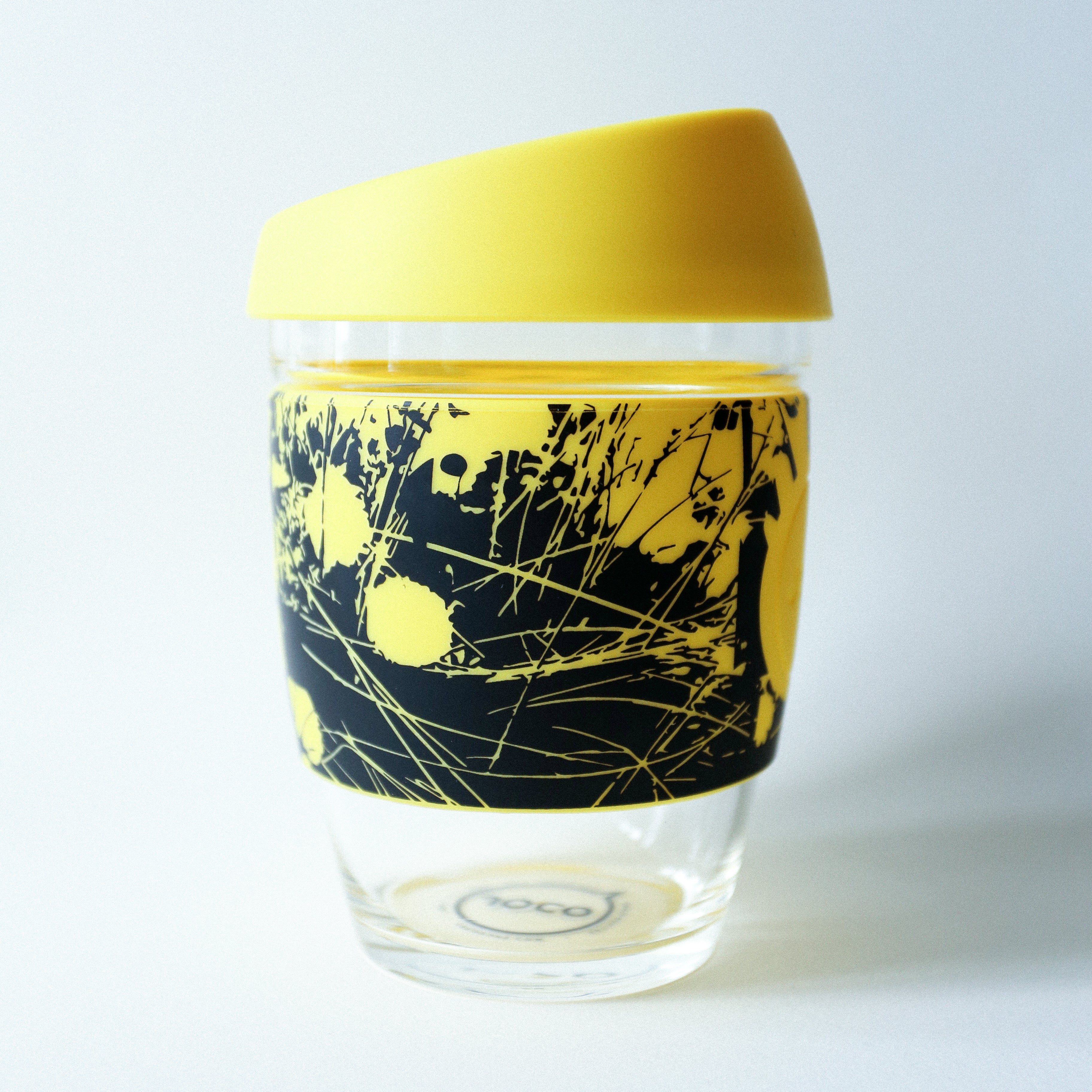 The Tandem x NUUFORM Joco Cup by vendor-unknown is a borosilicate glass cup with a bright yellow lid and a black-and-yellow sleeve featuring an abstract, nature-inspired design. This plastic-free reusable cup shines against a plain background.