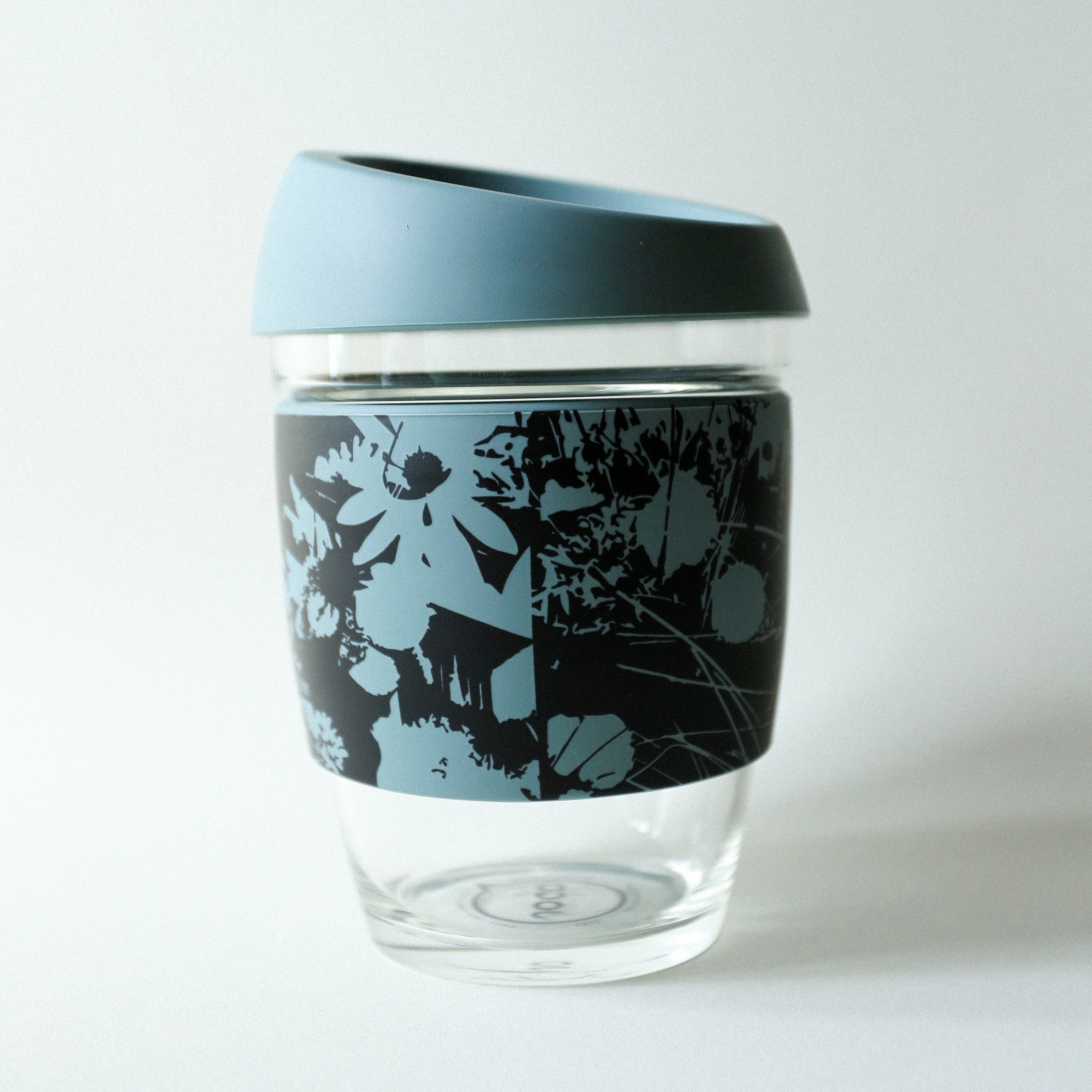 The Tandem x NUUFORM Joco Cup by vendor-unknown is a plastic-free, reusable borosilicate glass cup featuring a blue silicone lid and a black sleeve with light blue floral designs on white.