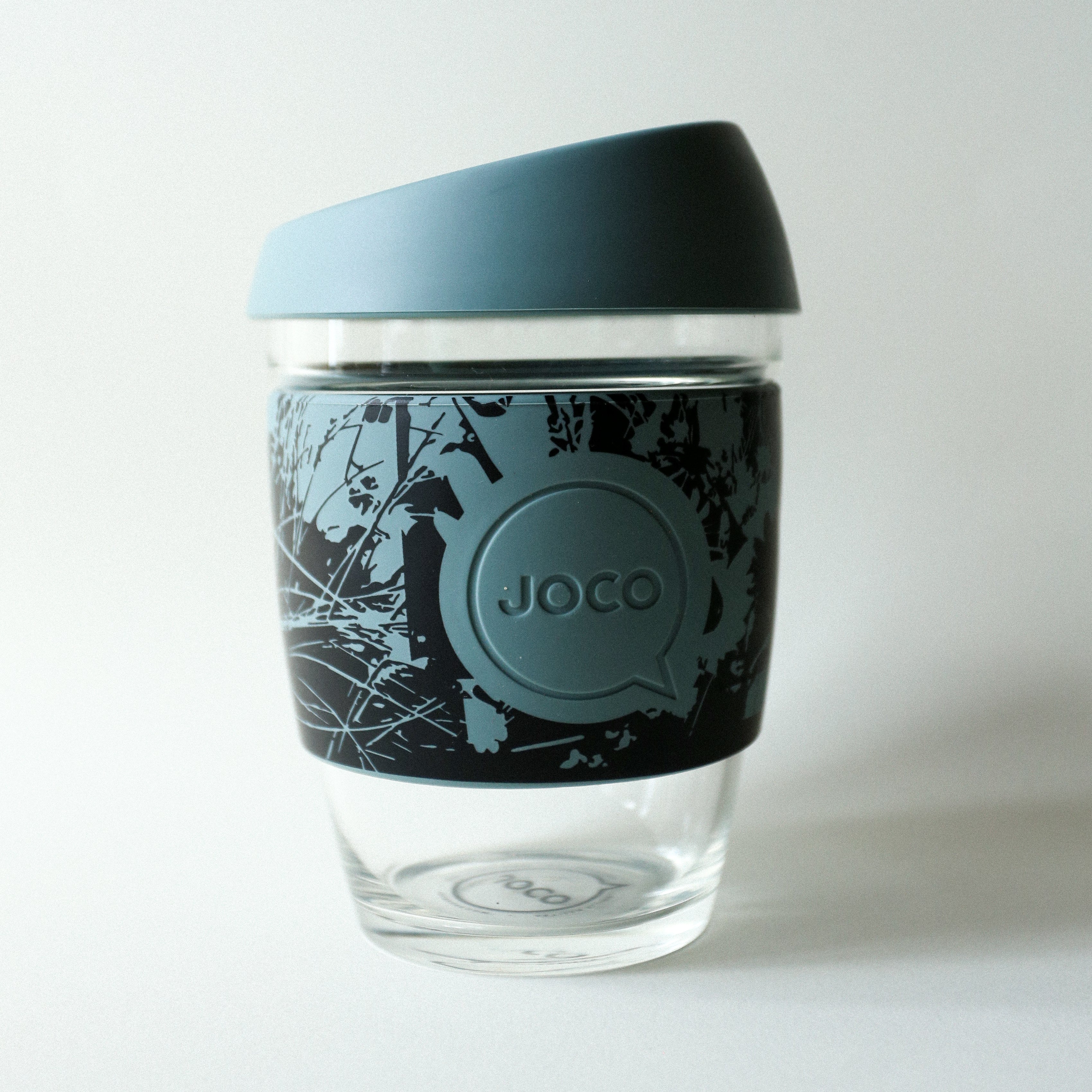 The Tandem x NUUFORM Joco Cup by vendor-unknown is a clear borosilicate glass cup with a blue nature-patterned silicone lid and sleeve, shown on a plain white background—an ideal plastic-free, reusable cup for daily use.