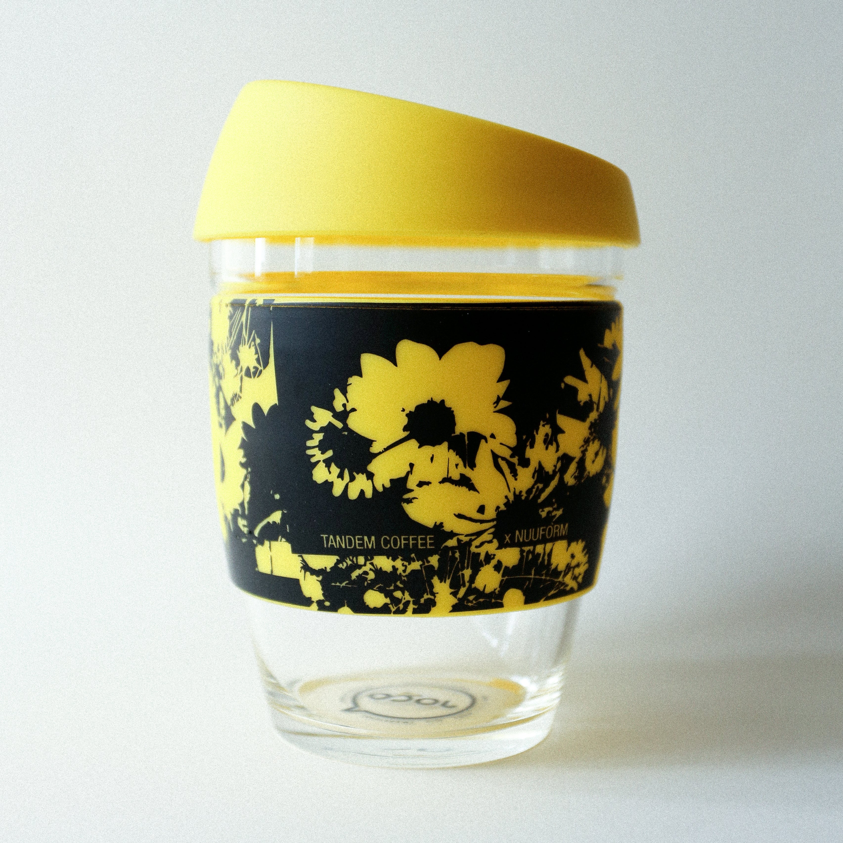 The Tandem x NUUFORM Joco Cup is a clear borosilicate glass cup with a yellow lid, wrapped with a black and yellow sunflower design and features TANDEM COFFEE x NUUFORM text on the sleeve. Brand: vendor-unknown.
