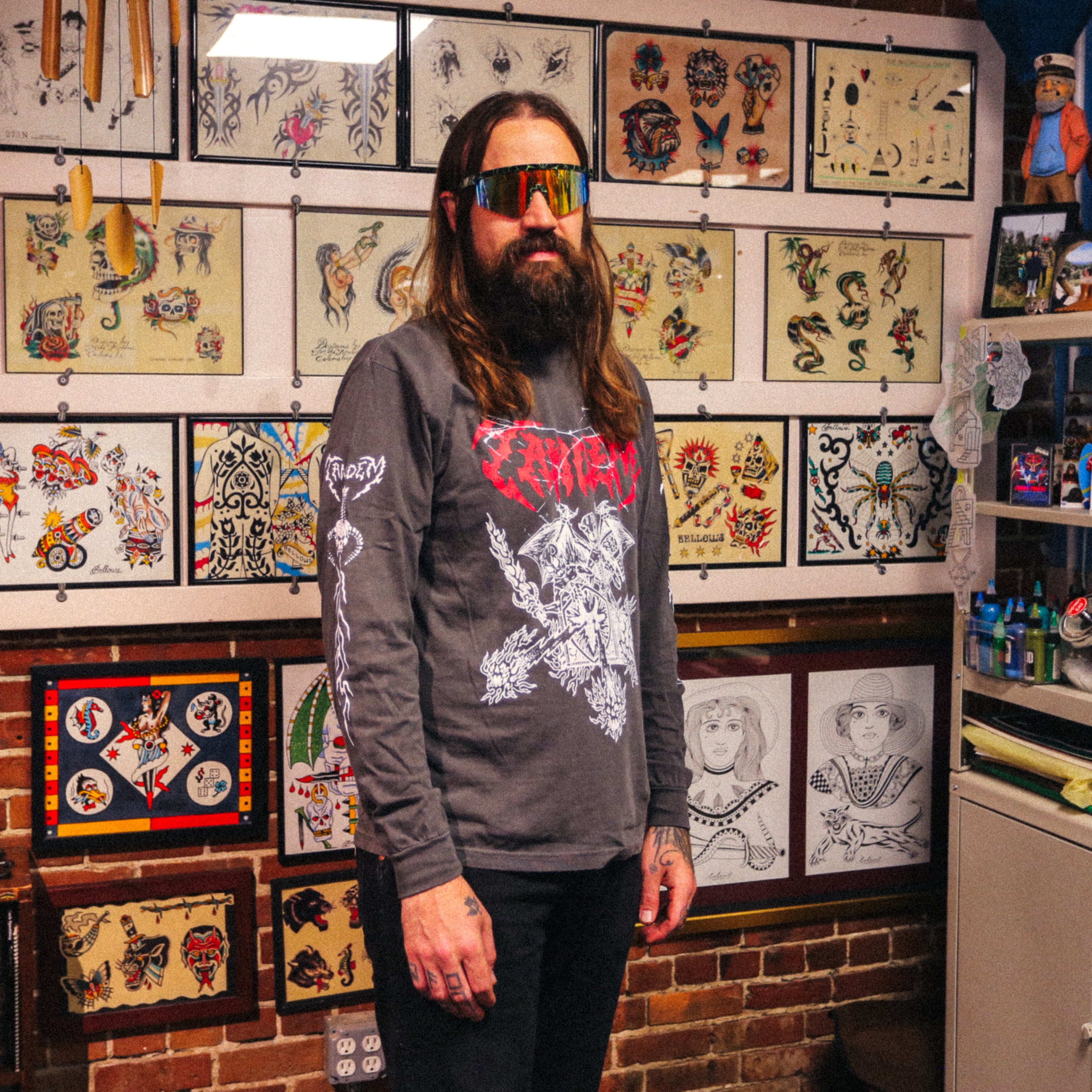 A person with long hair and sunglasses stands in Gold Star Tattoo studio, surrounded by framed tattoo art on brick and white walls. They wear a dark Tandem Coffee Roasters Tandem Metal Long Sleeve—a Comfort Color tee featuring bold white designs on the front and sleeves.