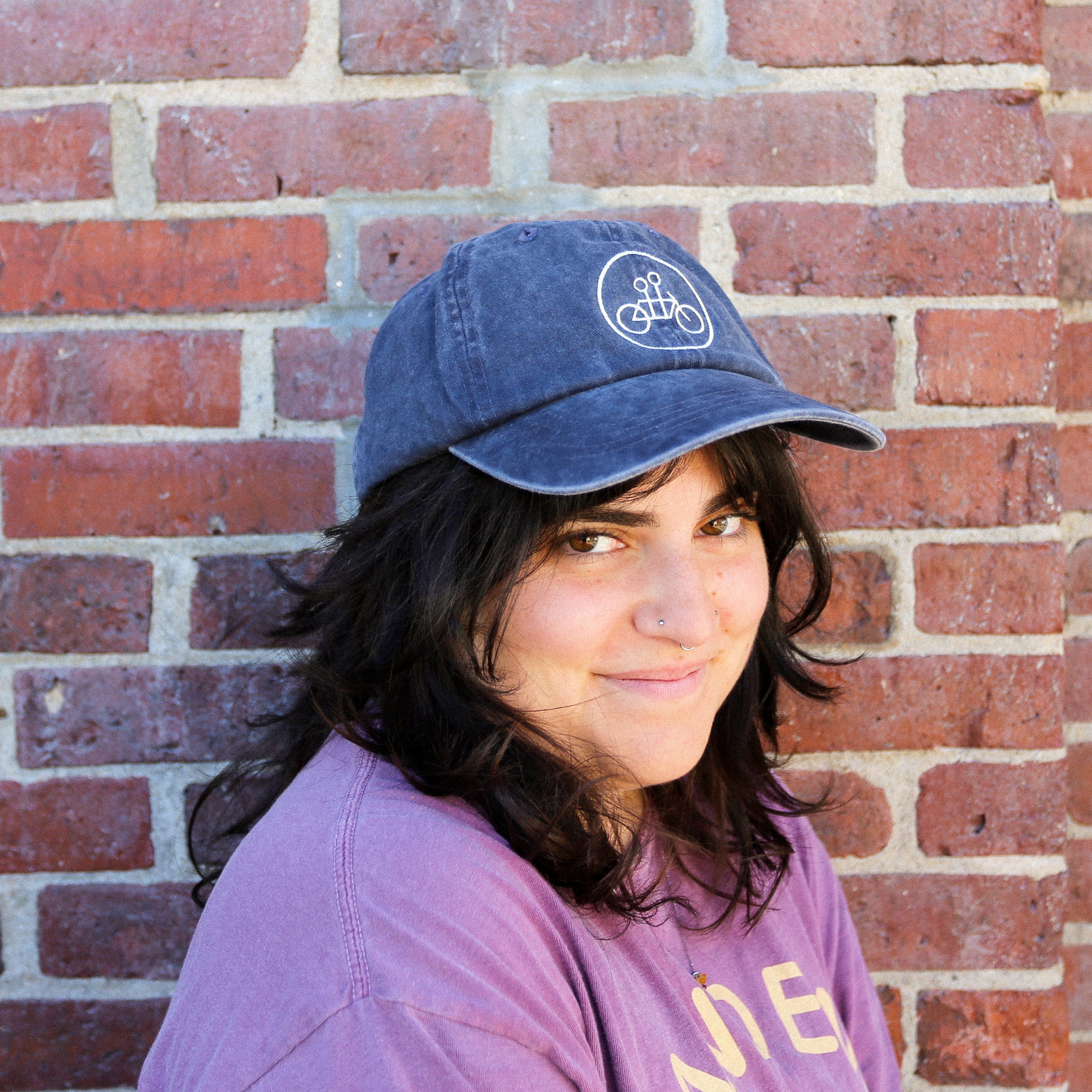 Person with long dark hair wearing a slate blue Circle Logo Dad Hat by Tandem Coffee Roasters and a purple shirt stands in front of a brick wall, smiling slightly. The cap features a white bicycle graphic on the front.