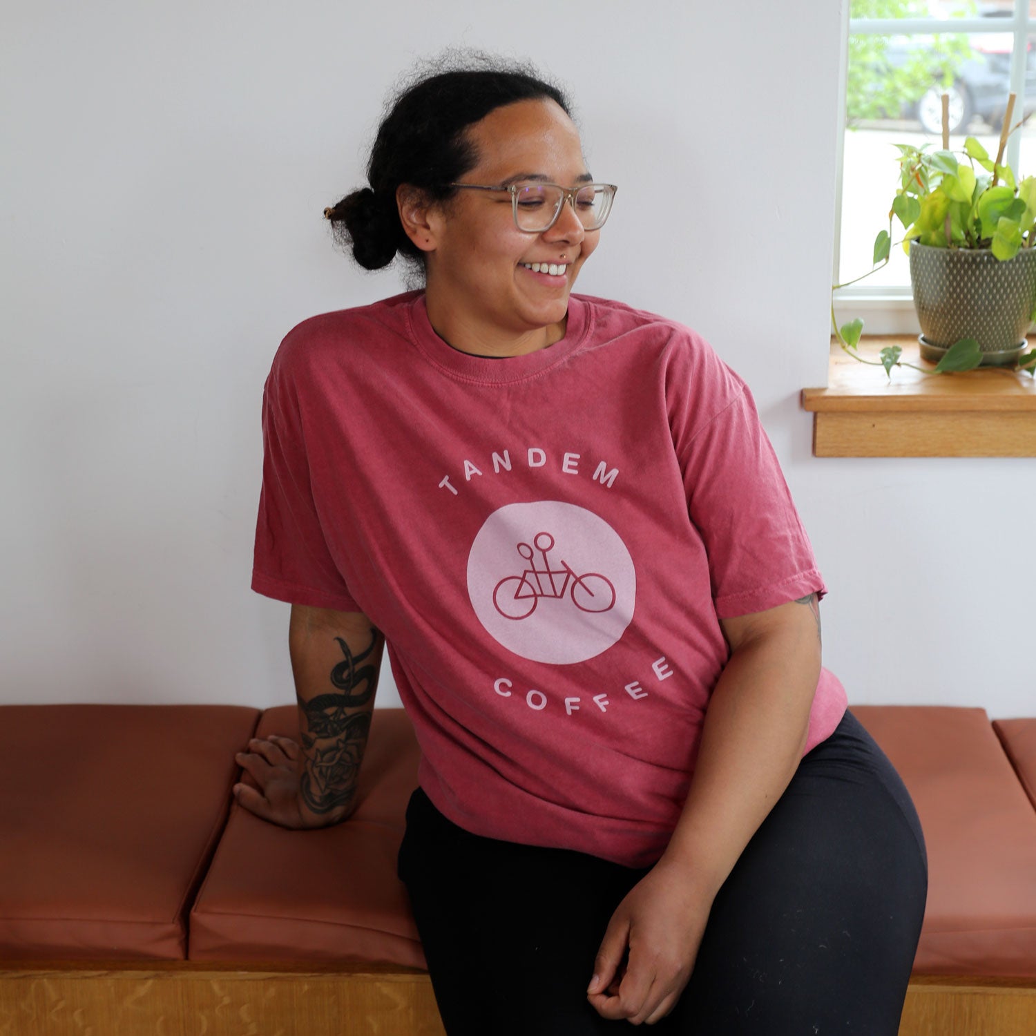 A person with glasses and a tattoo on their left forearm is smiling while sitting on a brown bench. They are wearing a Crimson Circle Logo Tee from Tandem Coffee Roasters with the text "Tandem Coffee" and an image of a tandem bicycle. A potted plant is on the windowsill behind them.