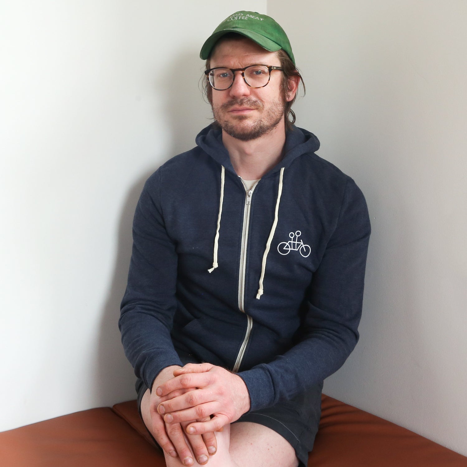 A man with glasses and a green cap sits on a brown bench against a plain white wall. He is wearing a cozy navy blue Letter Hoodie by Tandem Coffee Roasters with white drawstrings and a logo of a bicycle on the chest, along with gray shorts. His hands are clasped in his lap, and he is looking at the camera.