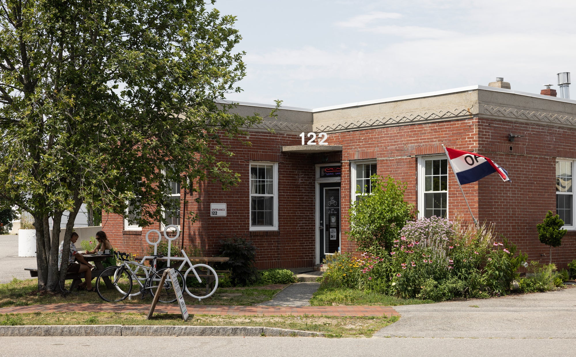 A quaint brick building with a patriotic flag, a bicycle sculpture in front, and a person seated at a table under a tree, likely enjoying a peaceful outdoor setting.