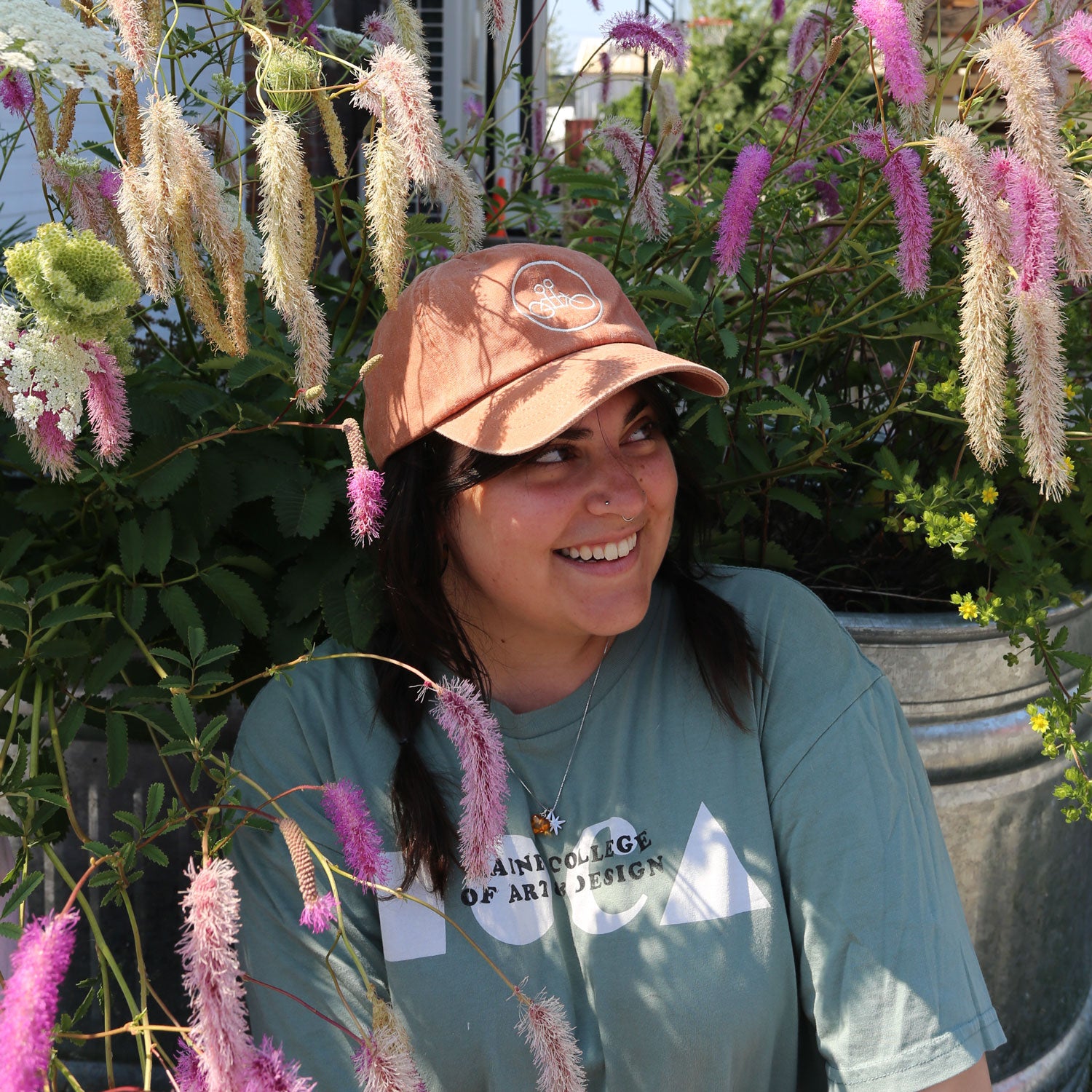 A person wearing a Tandem Coffee Roasters Rust Circle Logo Dad Hat and a green T-shirt smiles while sitting among tall, pink and white flowering plants on a sunny day.