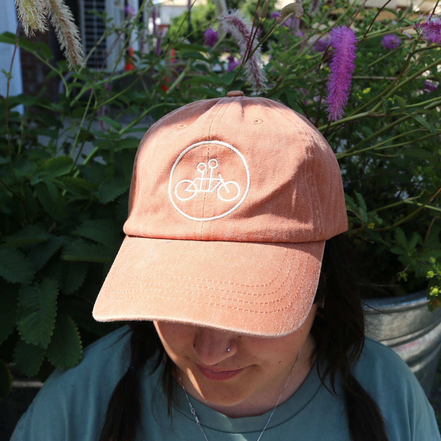 A person wears the Tandem Coffee Roasters Rust Circle Logo Dad Hat, an adjustable cotton cap with a white bicycle and scissors logo, standing among green plants with purple flowers. The hat sits low, partially obscuring their face.