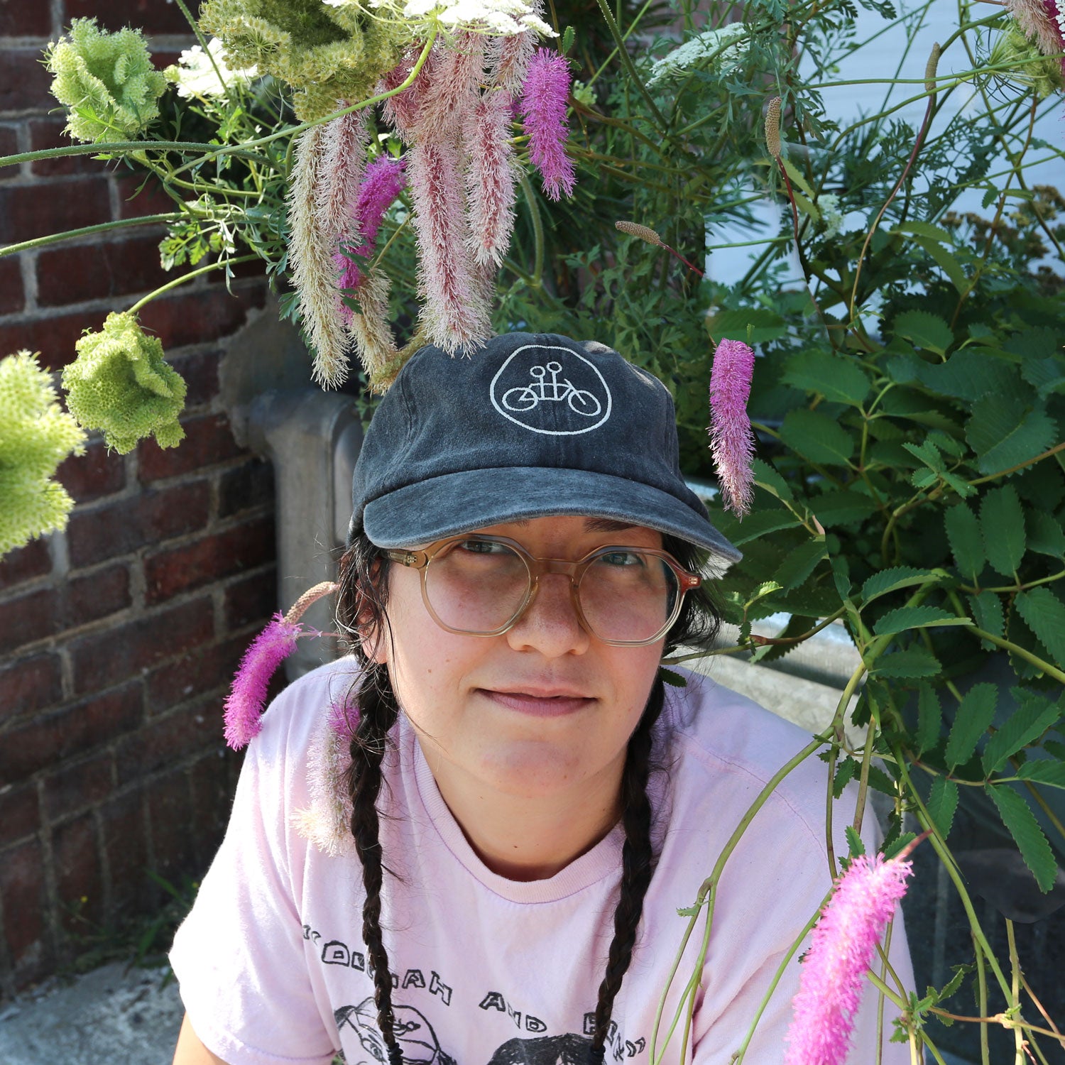A person with glasses and braids, wearing a light pink shirt and a Tandem Coffee Roasters Vintage Black Circle Logo Dad Hat, sits outdoors among pink and green flowers with a brick wall in the background.