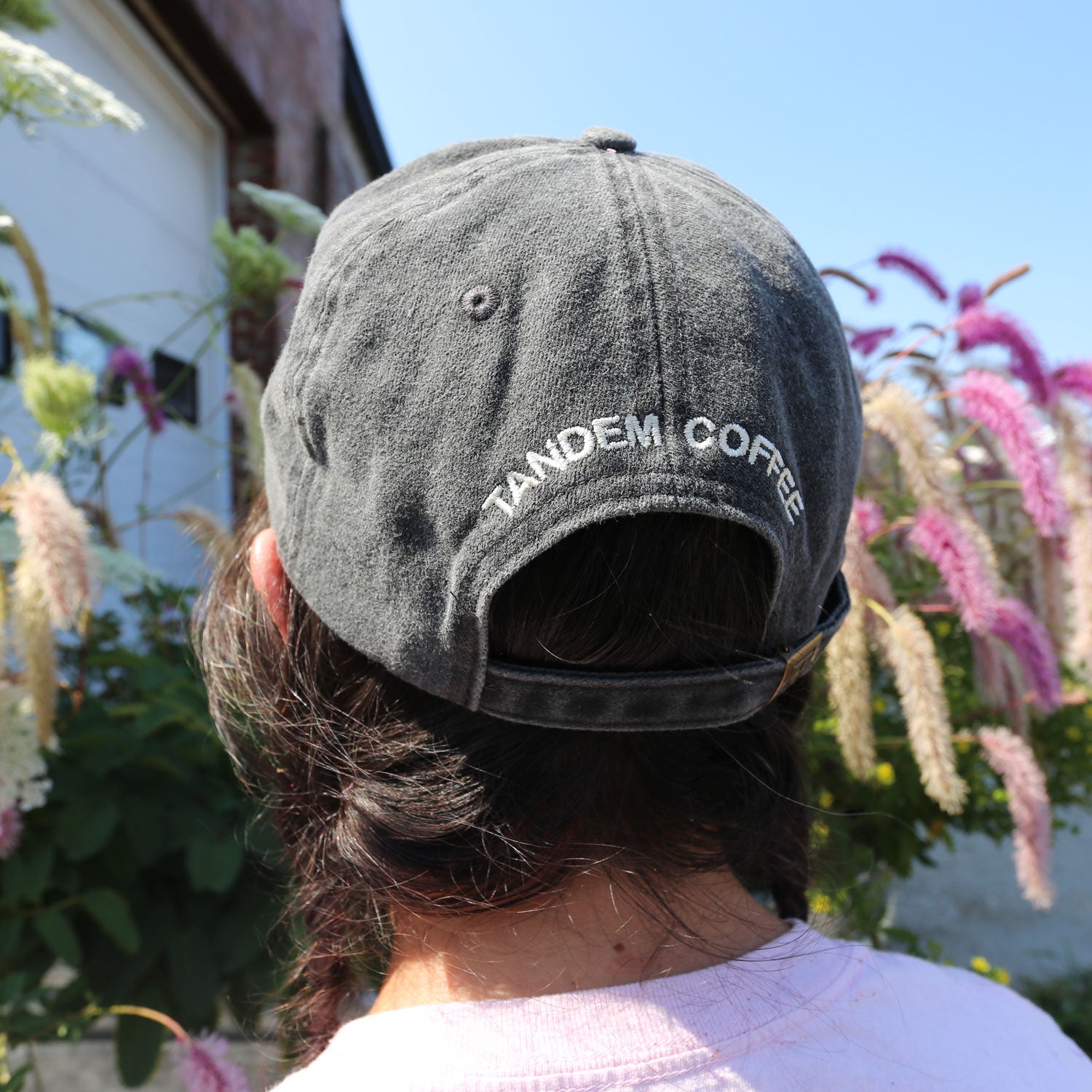 A person with brown hair wears a Vintage Black Circle Logo Dad Hat by Tandem Coffee Roasters, standing outdoors near flowering plants under a clear blue sky.