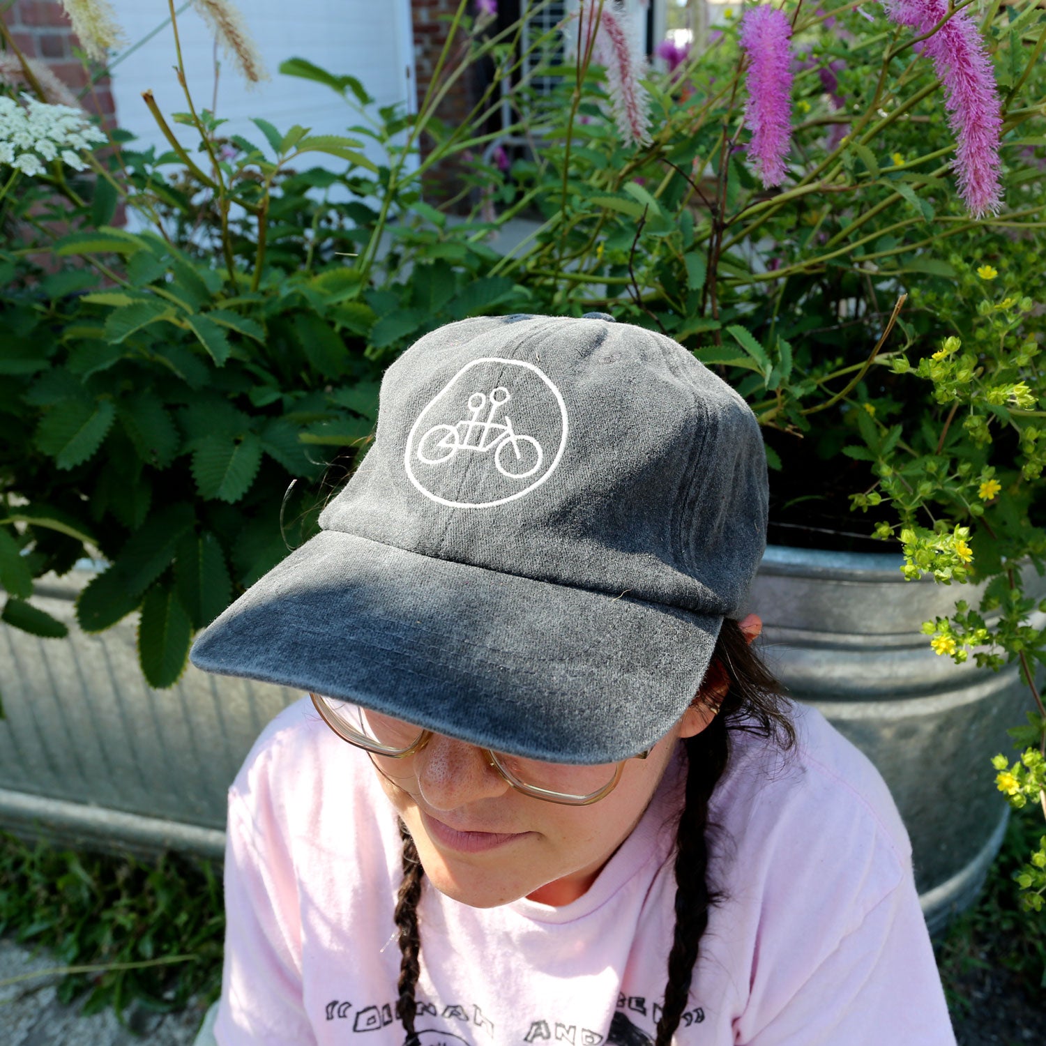 A person in glasses and a Vintage Black Circle Logo Dad Hat by Tandem Coffee Roasters—a gray, 100% brushed cotton embroidered hat with a bicycle logo—sits outdoors near green foliage and pink flowers, wearing a light pink shirt and braids.