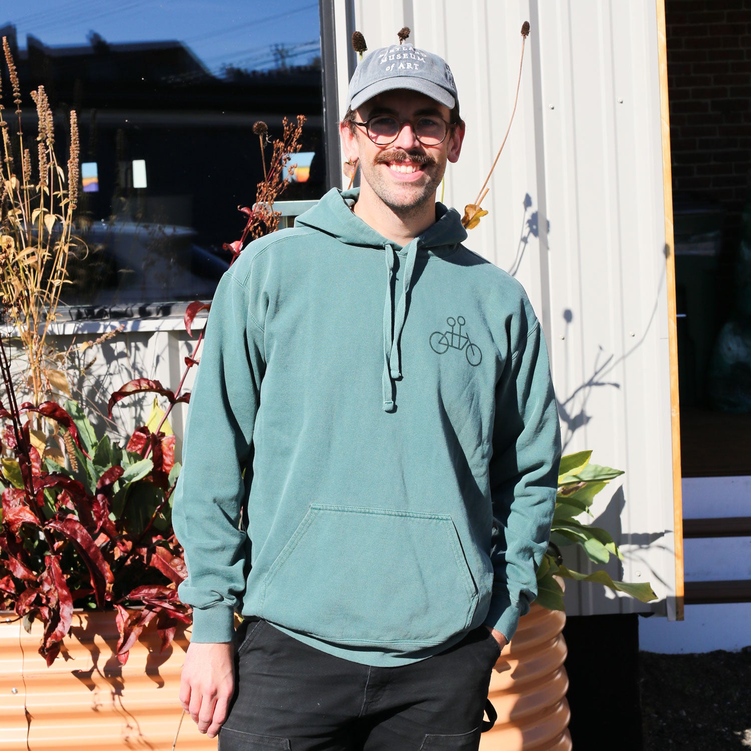 A smiling person with glasses, a mustache, and a light blue cap stands outside in sunlight wearing the Blue Spruce Tandem Hoodie from Tandem Coffee Roasters. Potted plants and a corrugated metal building are visible behind them.