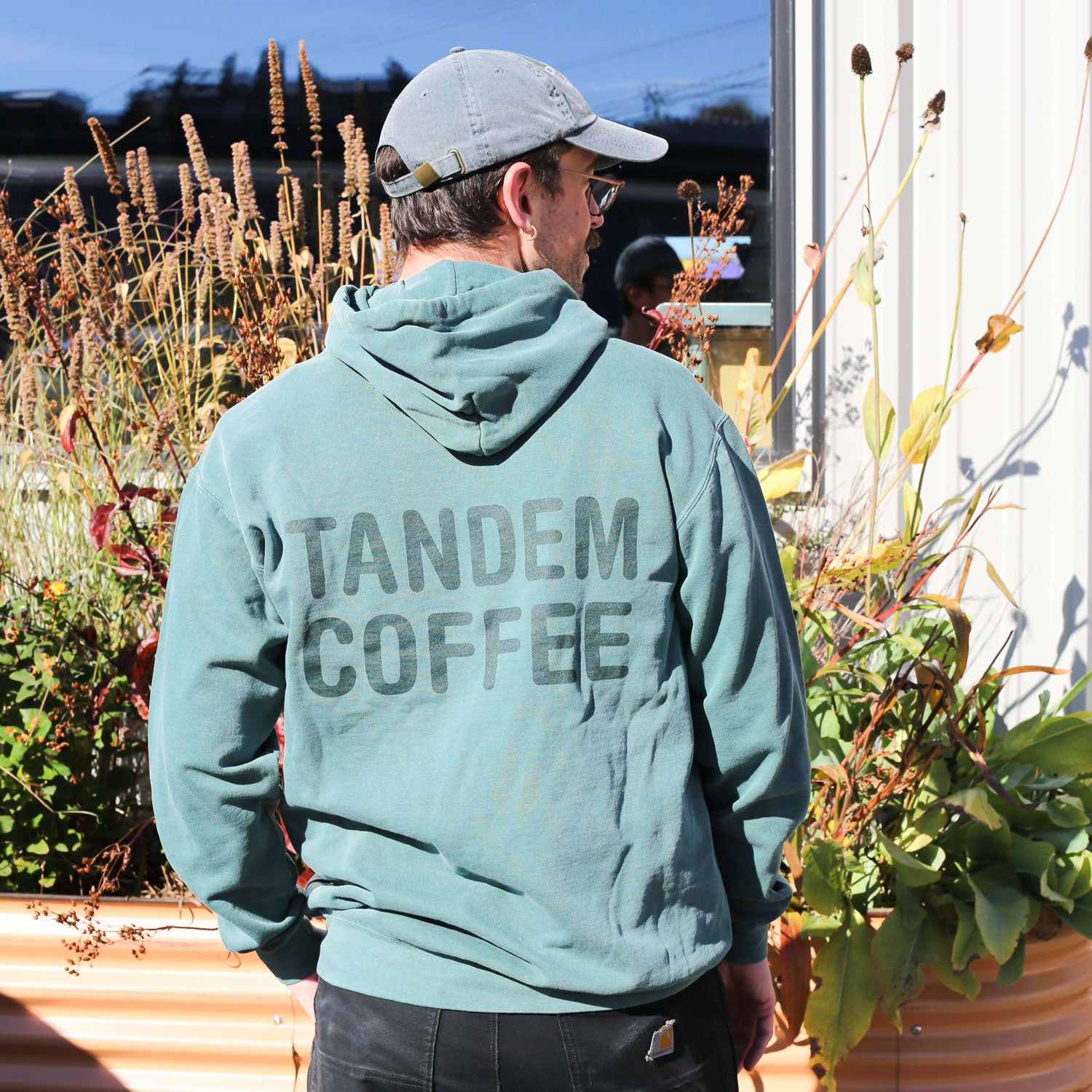 A person in a gray cap and a Blue Spruce Tandem Hoodie from Tandem Coffee Roasters, with "TANDEM COFFEE" on the back, stands outside near tall plants and a white building, facing away from the camera.