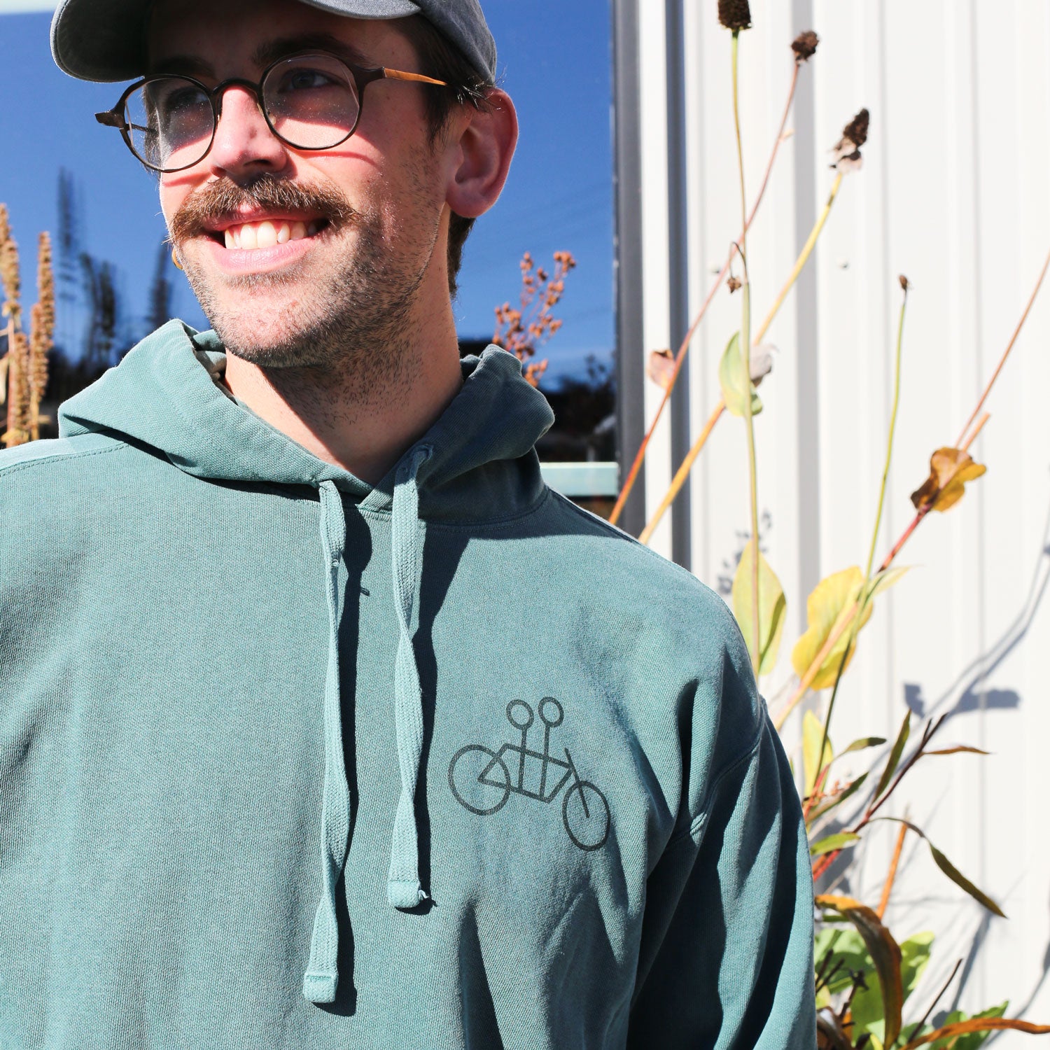 A smiling man with a mustache, glasses, and a gray cap wears the Blue Spruce Tandem Hoodie from Tandem Coffee Roasters. Featuring a simple bicycle graphic, he stands outdoors by tall plants and a metal building in gentle sunlight.