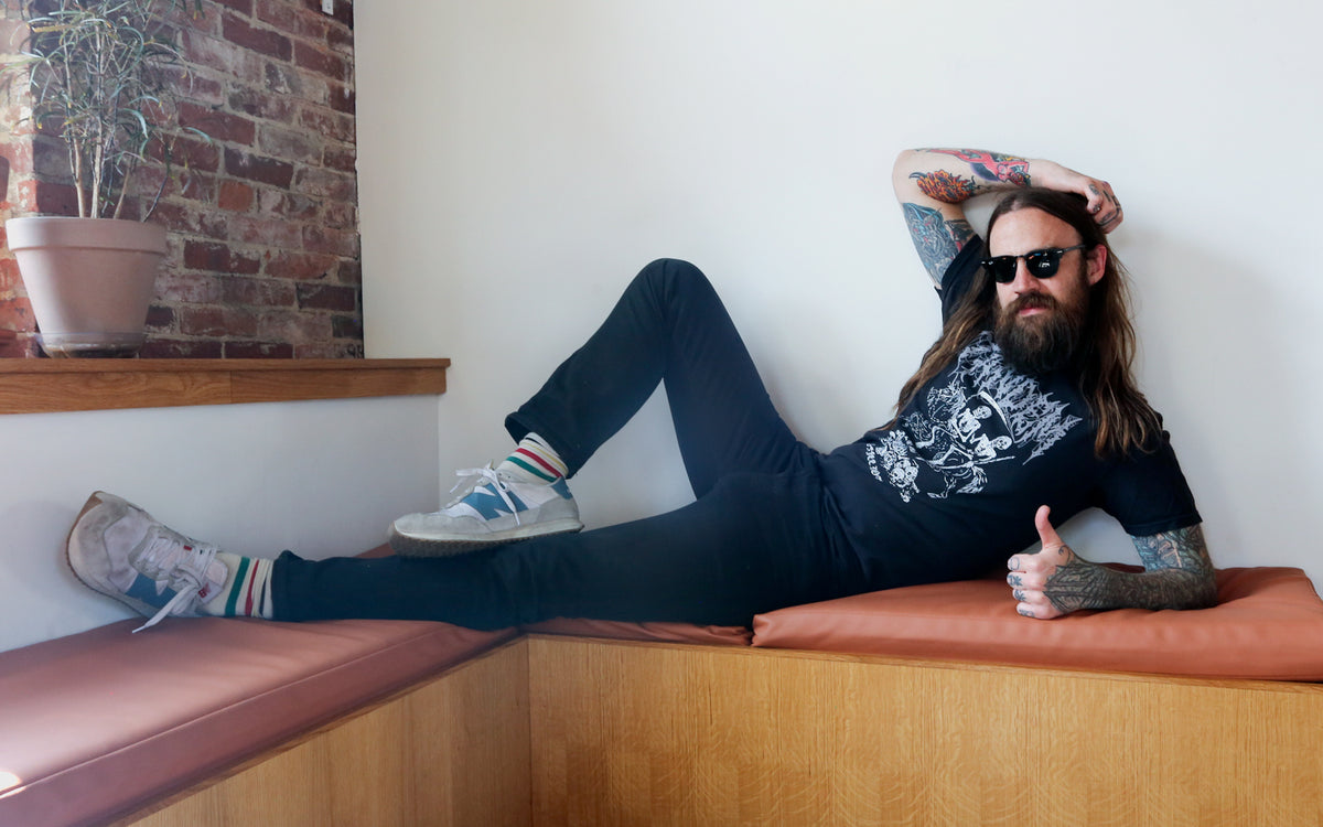 A man with long hair and tattoos reclines on an orange bench, wearing a black t-shirt, jeans, and sneakers, gesturing a thumbs up, with a brick wall and a potted plant in the background.