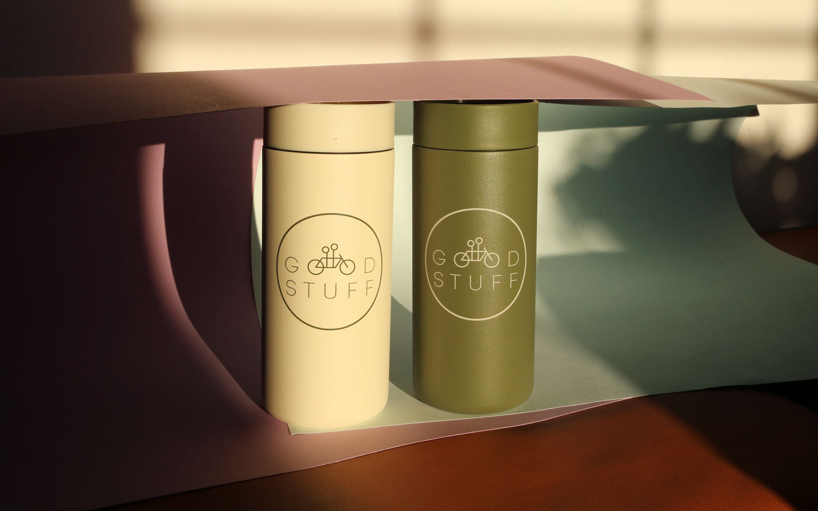 Two cylindrical containers labeled "good stuff" sit on a table, bathed in warm sunlight, casting soft shadows. one container is cream-colored and the other is olive green.