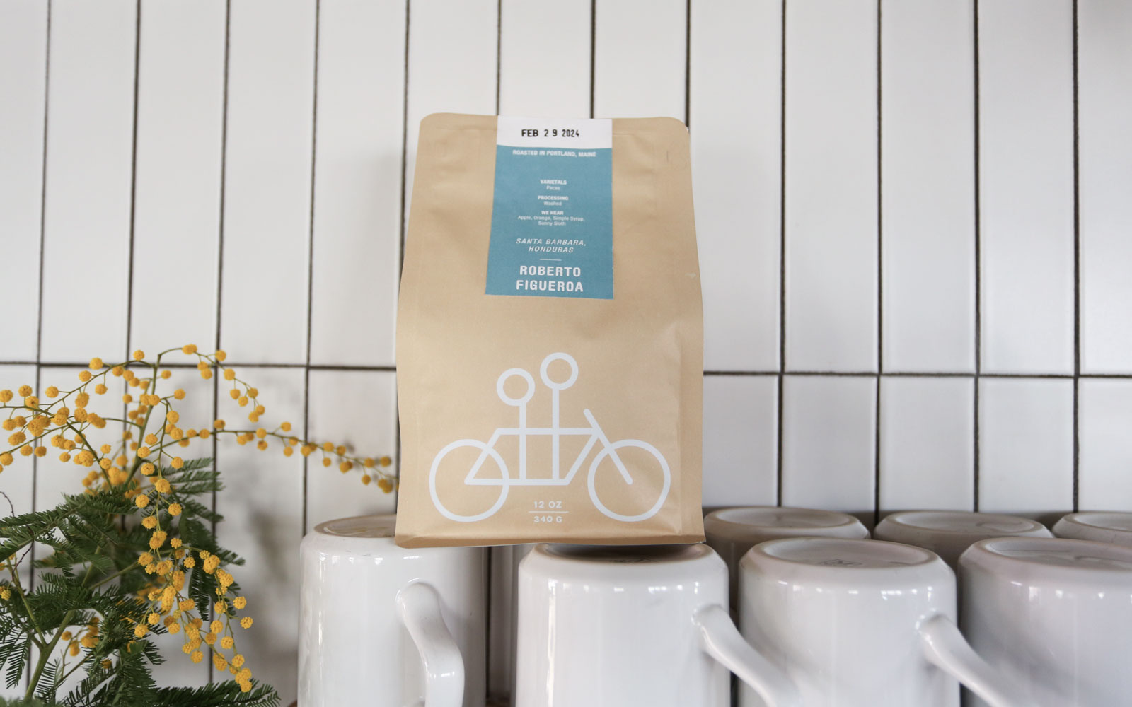 A coffee bag with a bicycle design stands on a white tiled countertop next to white mugs with yellow flowers in the background.