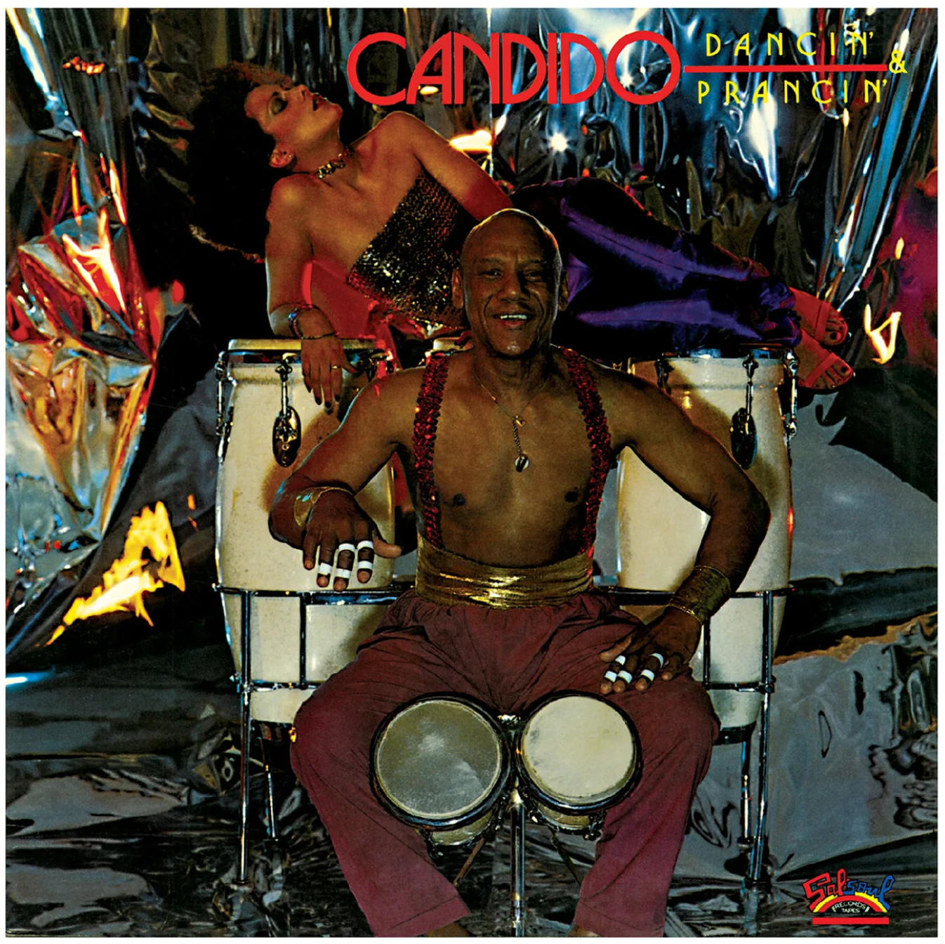 A man in red pants and suspenders plays conga drums, smiling, as a woman in a glittery top and purple skirt reclines on the drums. The metallic, colorful background features the "Candido - Dancin' and Prancin'" coffee by Tandem Coffee Roasters.