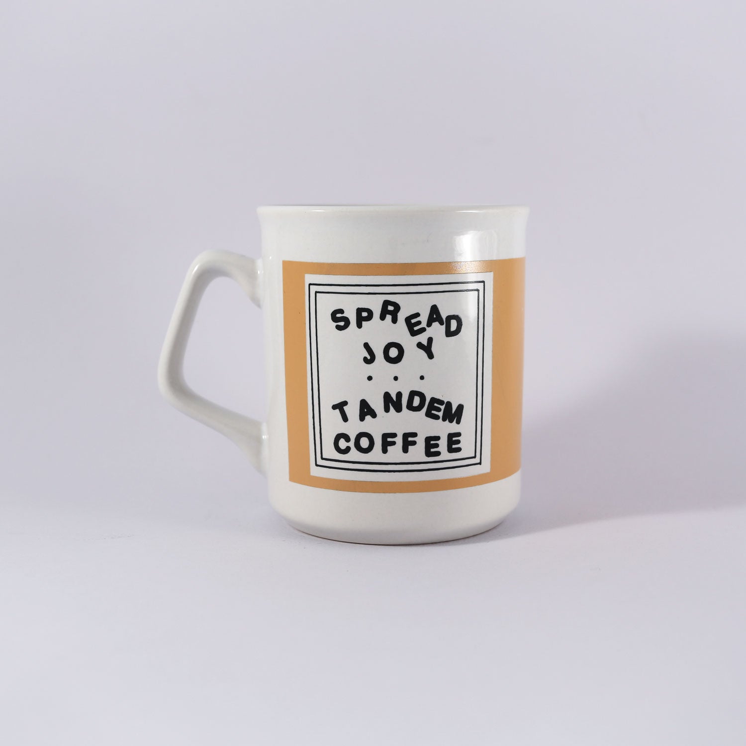 A white ceramic mug with a beige center band featuring the text "Kathy Heideman Spread Joy Mug" in a black square frame on a plain background, recommended for handwash only. Made by Tandem Coffee Roasters.