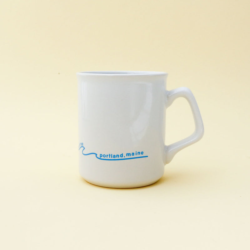 A classic white 11oz Sunshine Mug with a sun and a red bicycle design above a blue wave, set against a pale yellow background by Tandem Coffee Roasters.