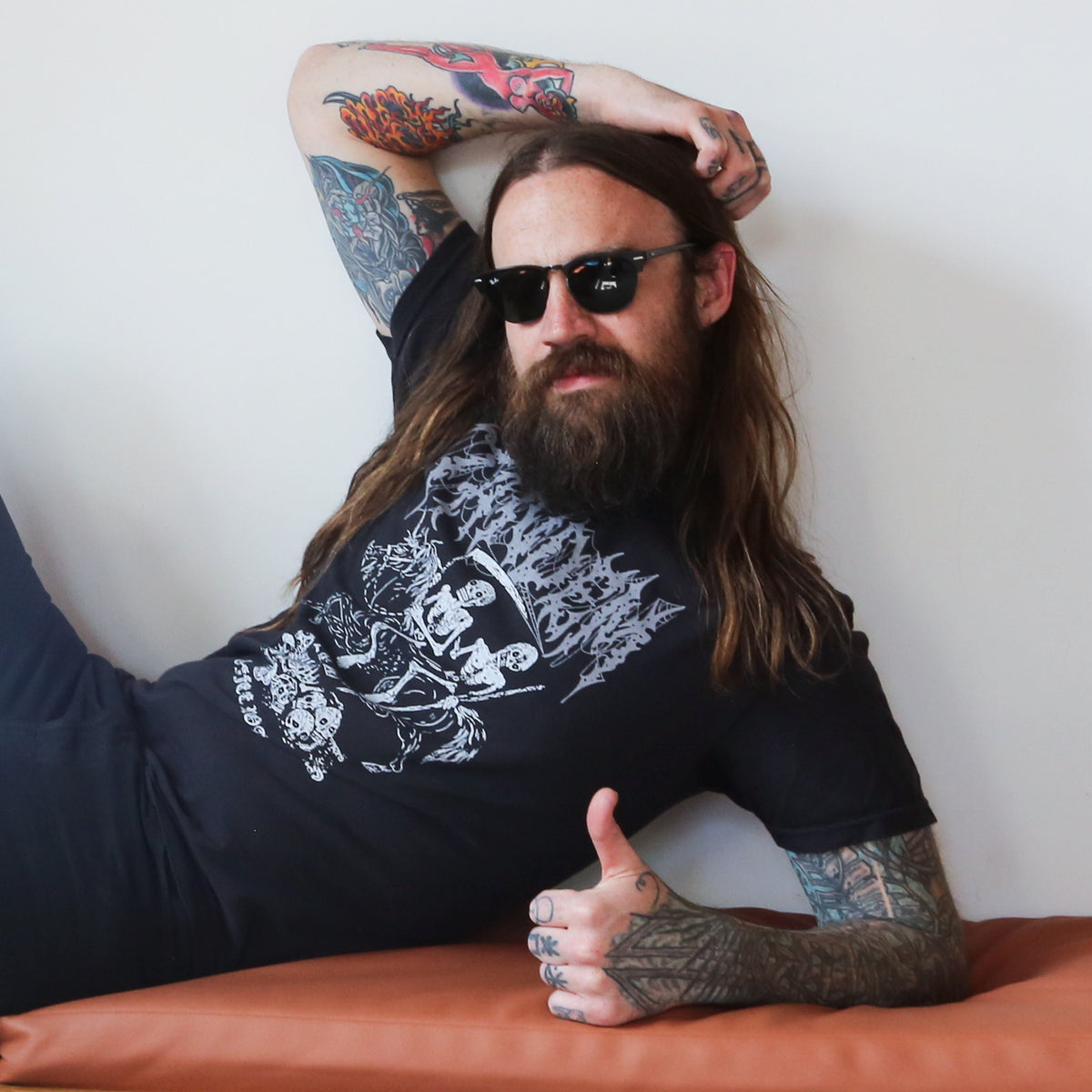 A man with long hair and tattoos wearing sunglasses and a Tandem Coffee Roasters Gold Star Tattoo graphic t-shirt gives a thumbs-up while reclining on an orange bench.