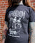 A person with tattooed arms wears a black Tandem Coffee Roasters tee with a white graphic of skeletons and text. They stand facing away, against a brick wall background.