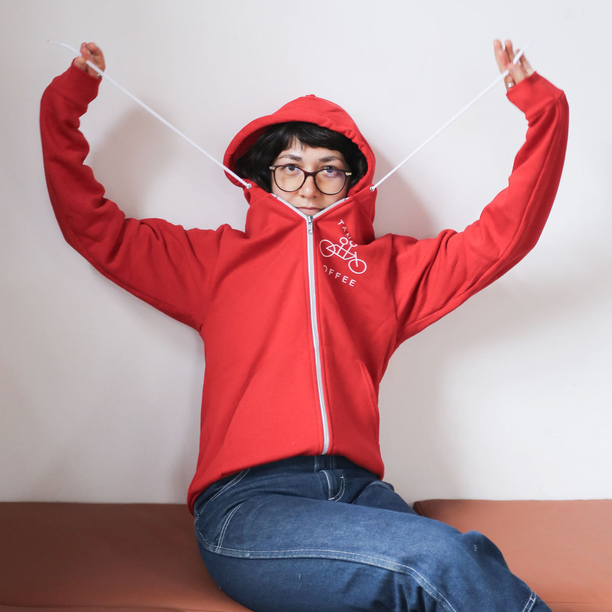 A person in a Tandem Coffee Roasters red zip-up hoodie with a bicycle logo sits on a bench, playfully pulling the hoodie's drawstrings while wearing glasses and a hood over their head.