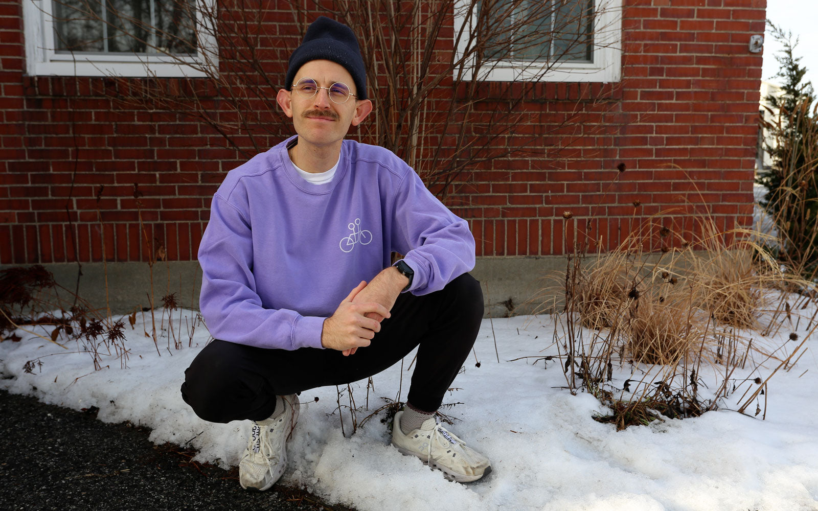 A man squatting by a snow-covered ground outside a brick building, wearing a purple sweatshirt, blue beanie, glasses, and sneakers, smiling at the camera.
