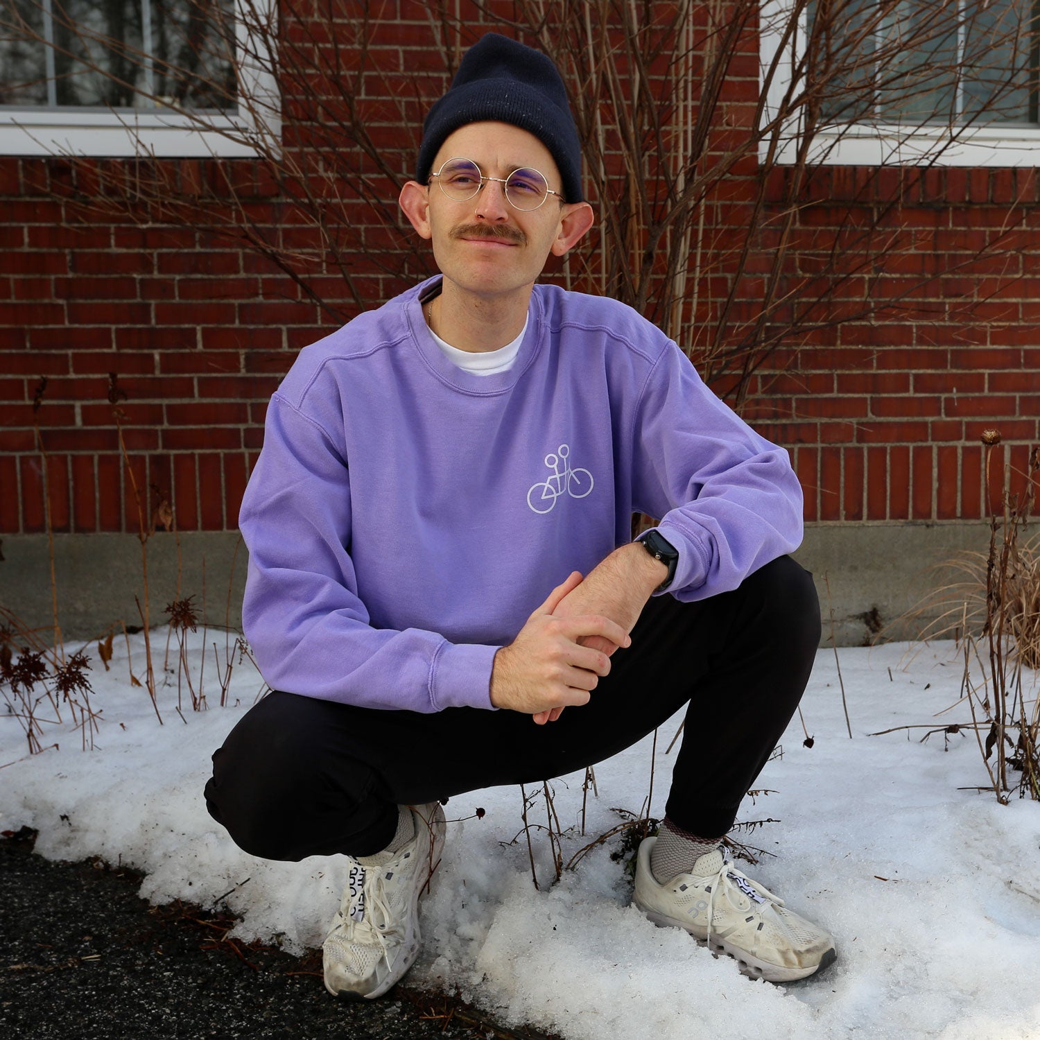 A person squats on a snowy path before a brick building, wearing the cozy purple Tandem Crewneck Sweatshirt by Tandem Coffee Roasters, black pants, white sneakers, a dark beanie, and glasses. Sporting a mustache and wristwatch, they smile at the camera.