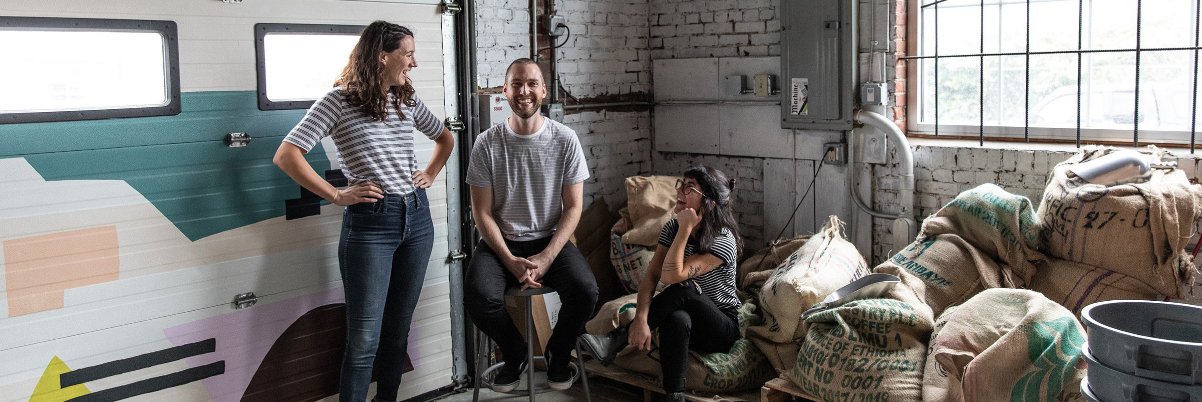 Three people in a casual setting inside a rustic, brick-walled room. One person stands with a hand on their hip, another sits on a stool smiling, and the third person sits on a stack of sacks, covering their face playfully. They seem to be enjoying some Tandem Coffee from Portland, Maine together.