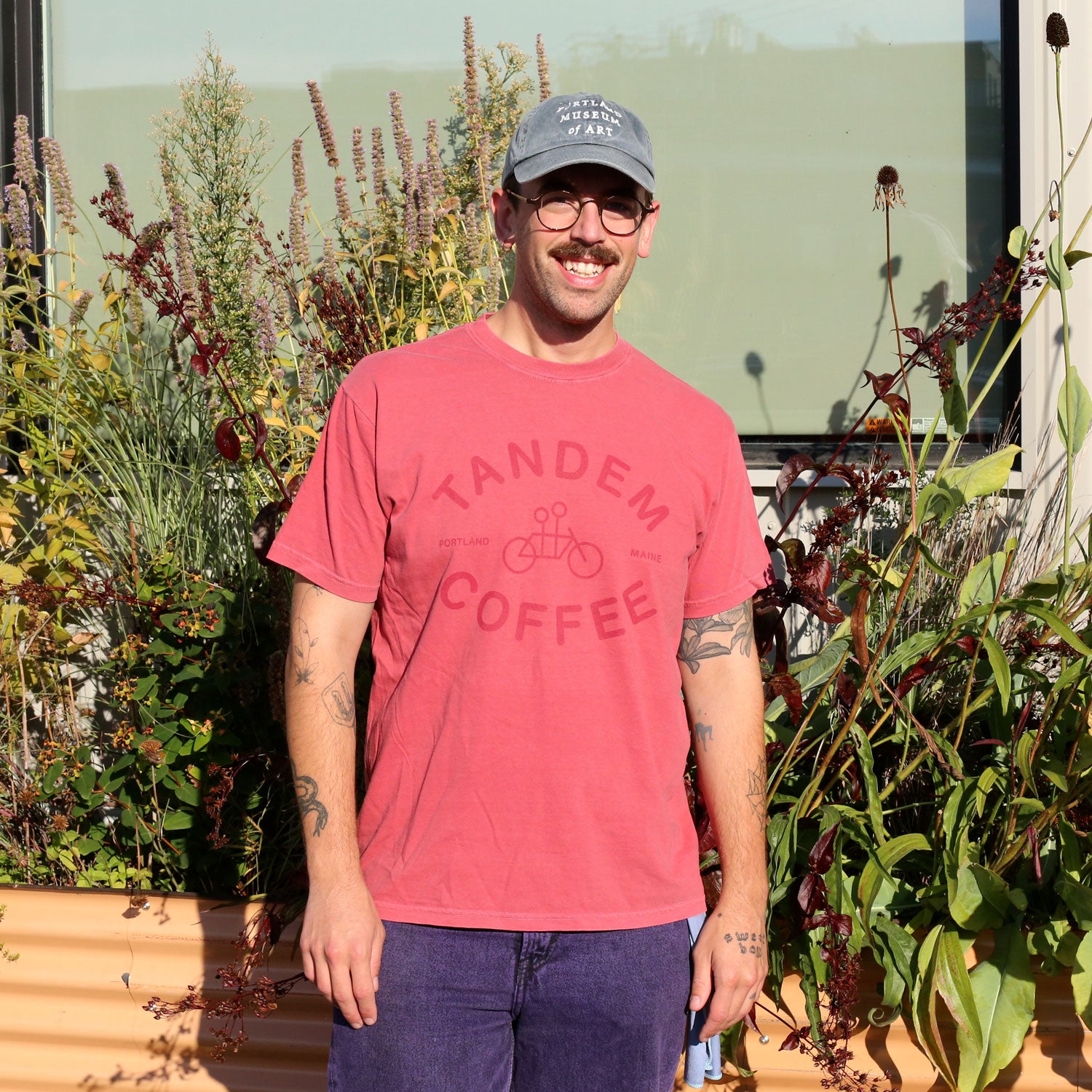 A man in a gray cap and glasses smiles outside by tall plants, wearing the Tandem Coffee Roasters Crimson Logo Tee. The 100% cotton Comfort Colors shirt enhances his relaxed, cheerful vibe.