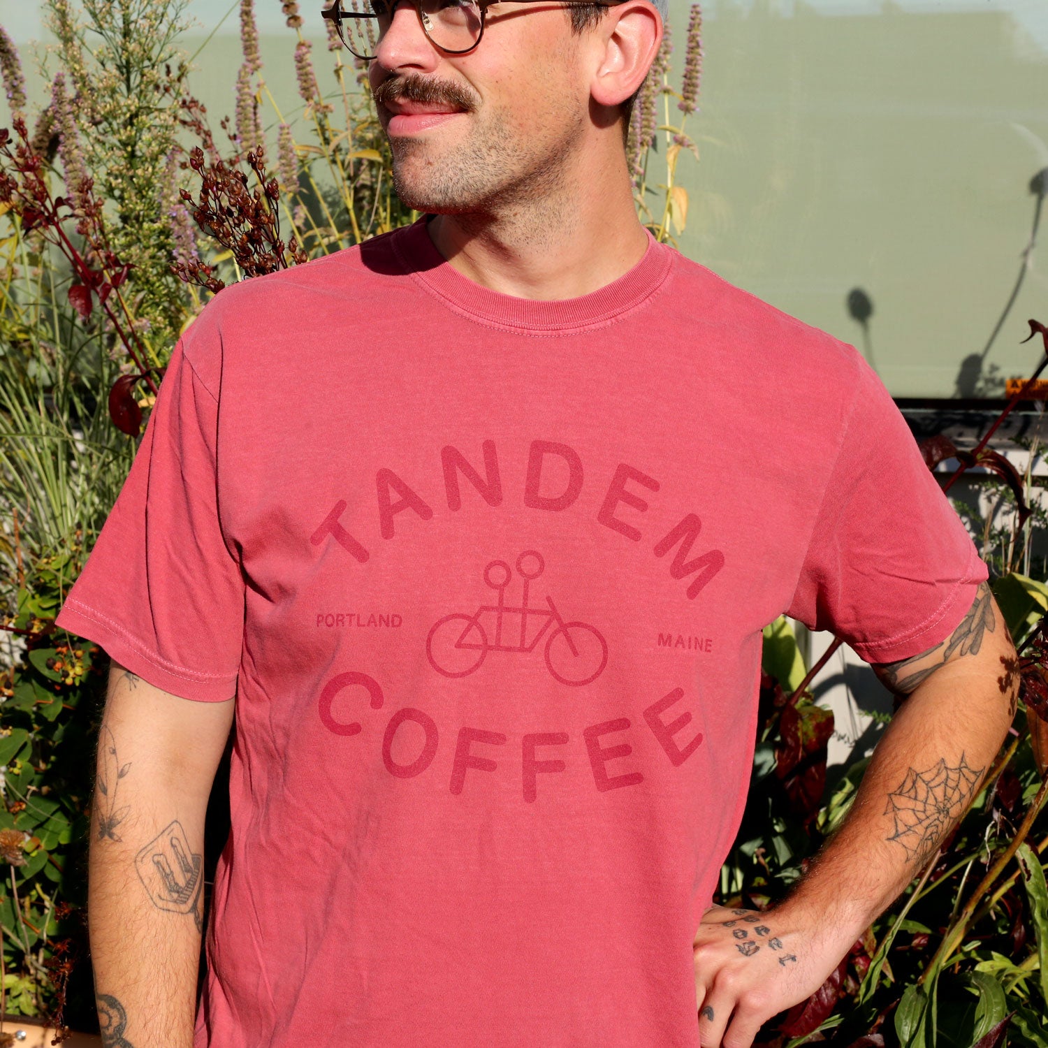 A tattooed person with a mustache and glasses wears the Crimson Logo Tee by Tandem Coffee Roasters, made from 100% cotton, while standing outdoors in sunlight among green plants and flowers.