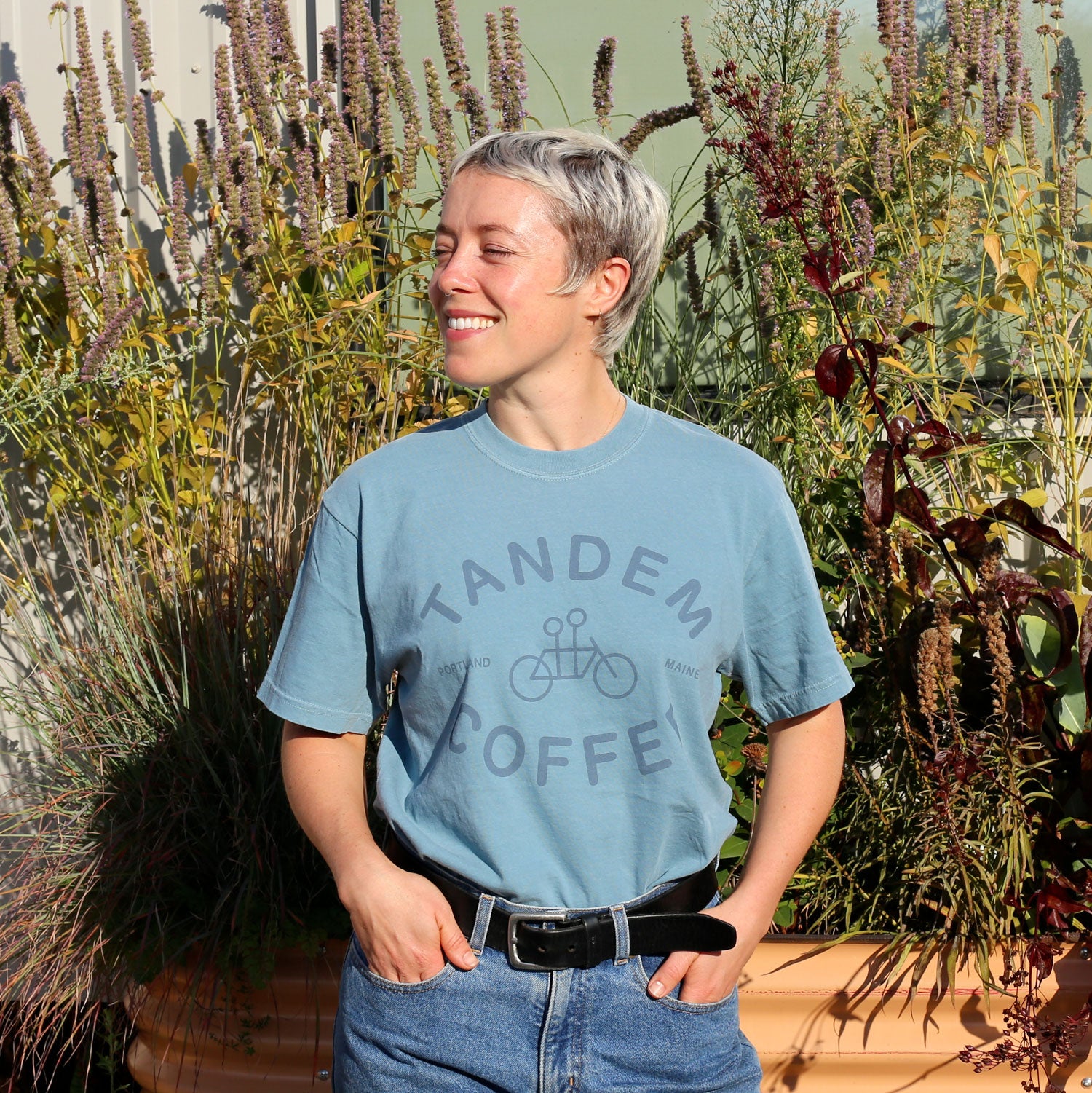 A smiling person with short blond hair stands outside in front of tall plants, wearing a Tandem Coffee Roasters Ice Blue Logo Tee and blue jeans with a black belt, one hand tucked casually in their pocket.