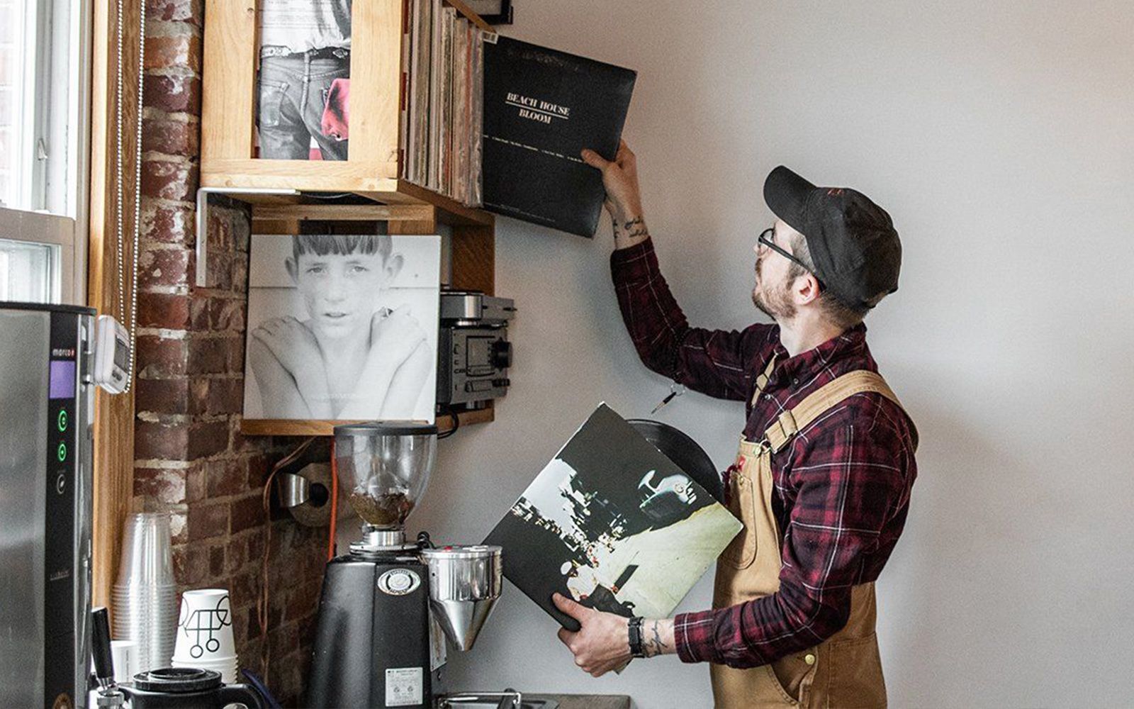 A man in a hat and suspenders is selecting vinyl records from a shelf in a cozy brick-walled room, with a coffee grinder visible on the left.