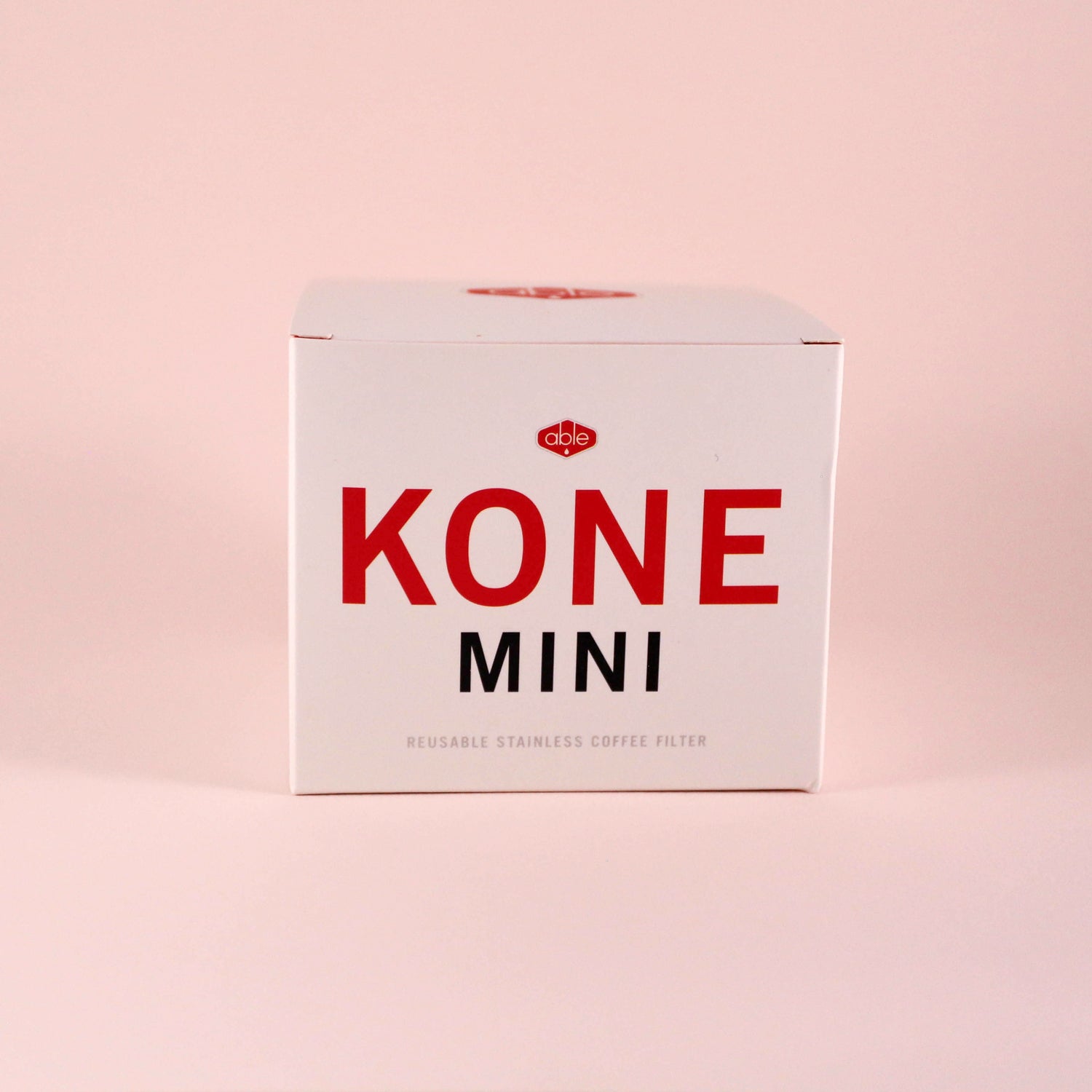 A gray, cone-shaped reusable stainless steel filter titled "Tandem Coffee Kone Mini" leaning against its packaging box on a pink background.