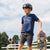 A young skateboarder in a helmet and 100% cotton Tandem Kids Tee stands confidently with a skateboard, looking off into the distance, with another person in the background at a skate park.