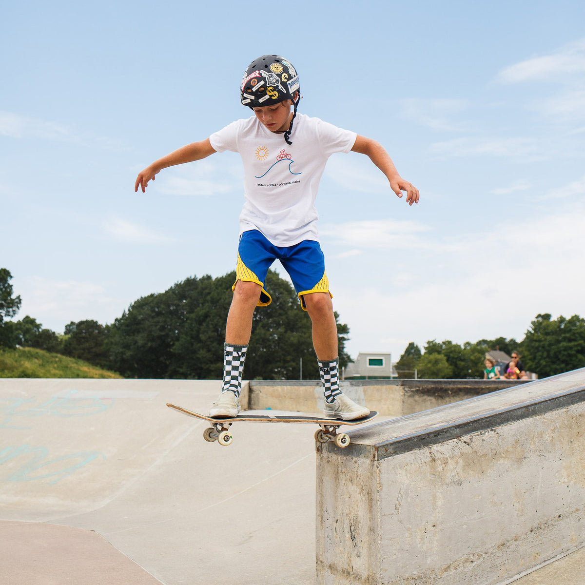 A young skateboarder, wearing a Tandem Kids Sun Tee, blue shorts, and knee-high socks, performs a trick at the edge of a skatepark ramp, focused and balanced on his board.