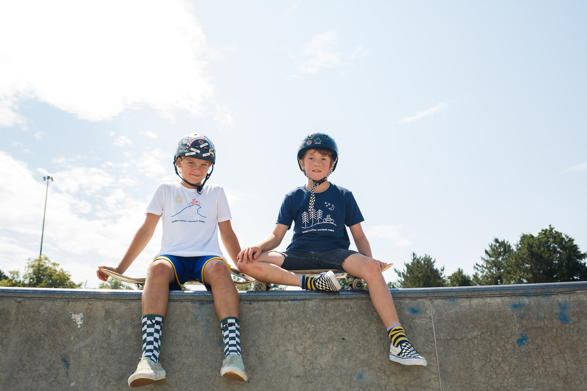 Two young boys wearing Tandem Kids Tandem Moon tees and helmets sit on the edge of a skatepark ramp on a sunny day, smiling and looking relaxed.