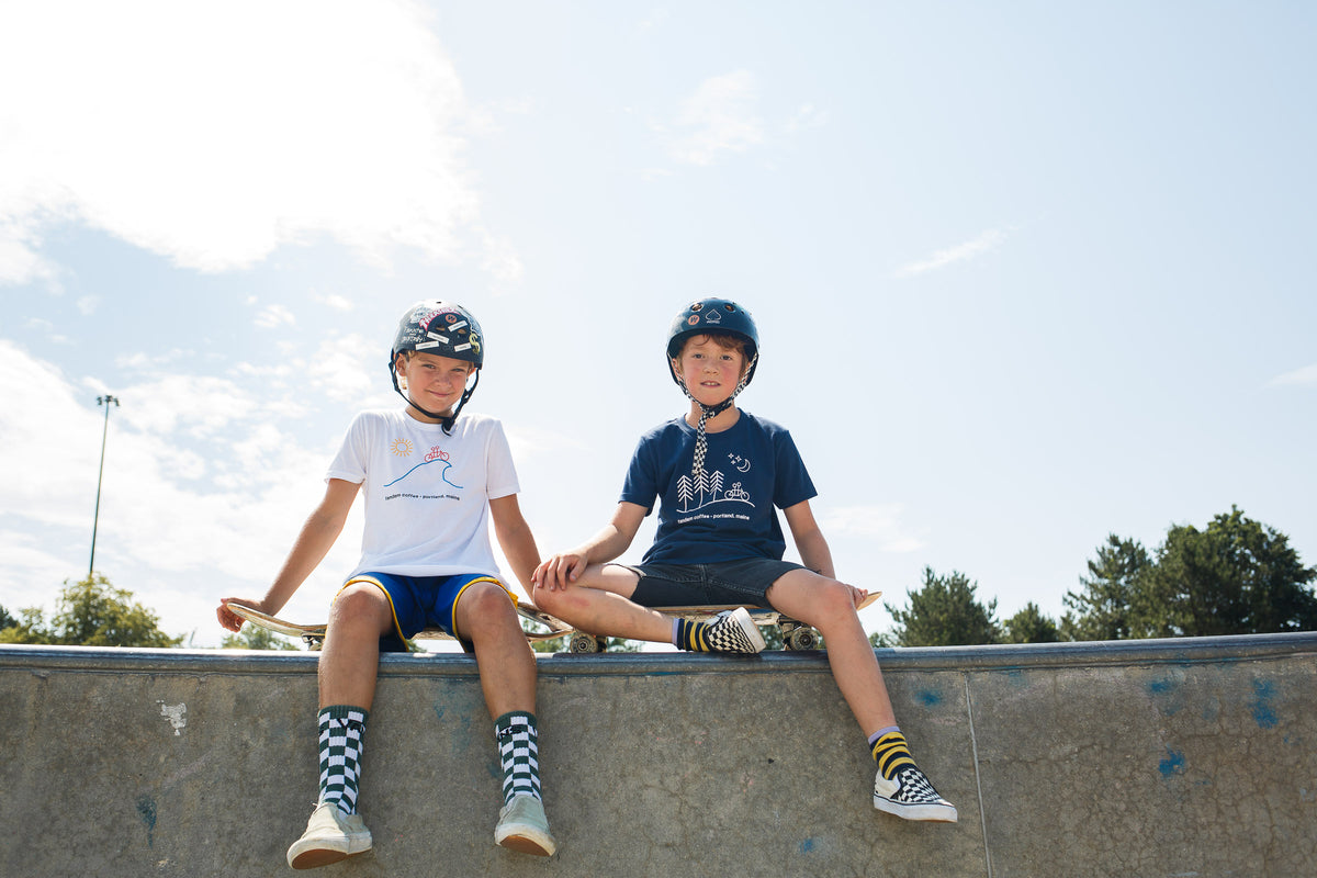 Two young boys wearing helmets and Tandem Kids Tandem Sun tees sit on the edge of a skateboard ramp under a clear blue sky, smiling and looking relaxed.